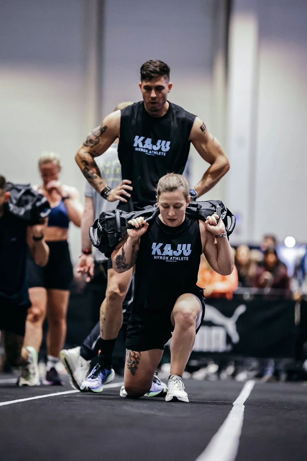Zwei Sportler beim Krafttraining im Fitnessstudio, beide tragen schwarze Shirts mit der Aufschrift 'KAJU HYROX ATHLETE', eine Person kniet mit einer Gewichtsriegel auf den Schultern, die andere Person steht dahinter mit Händen in den Hüften, im Hintergrund sind andere Sportler und ein Puma-Logo sichtbar.