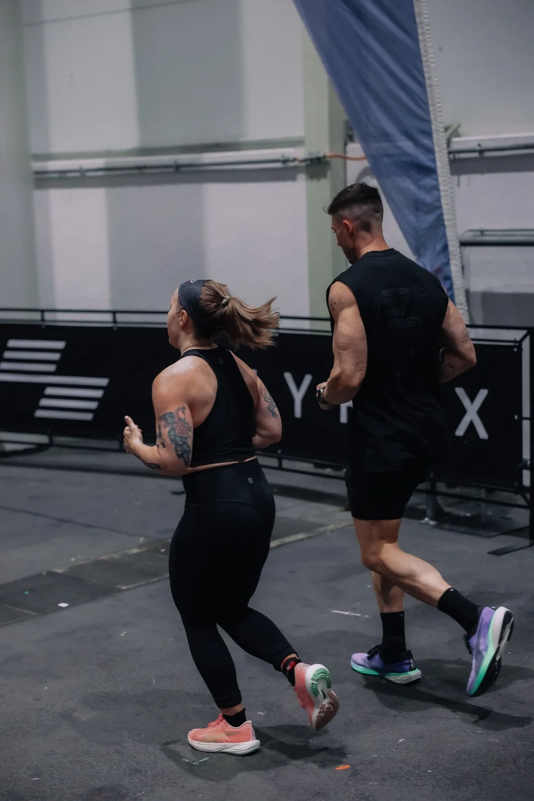 Zwei Menschen laufen im Fitnessstudio, einer ist eine Frau mit Tattoos und einer ist ein Mann, beide tragen Sportkleidung und laufen auf einem Laufband.