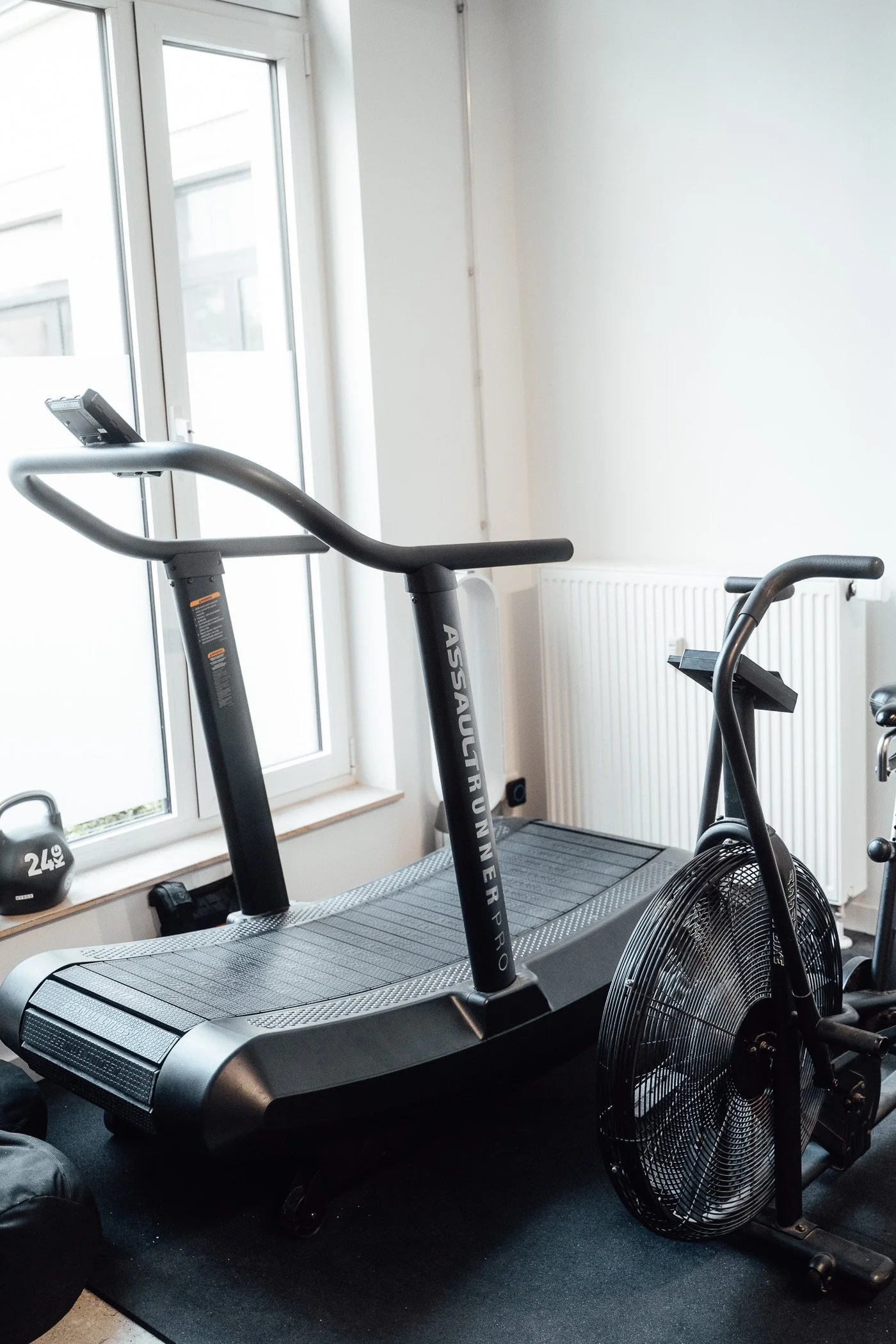 Ein Heim-Fitnessstudio mit Laufband und stationärem Fahrrad vor einem Fenster und einer weißen Wand.