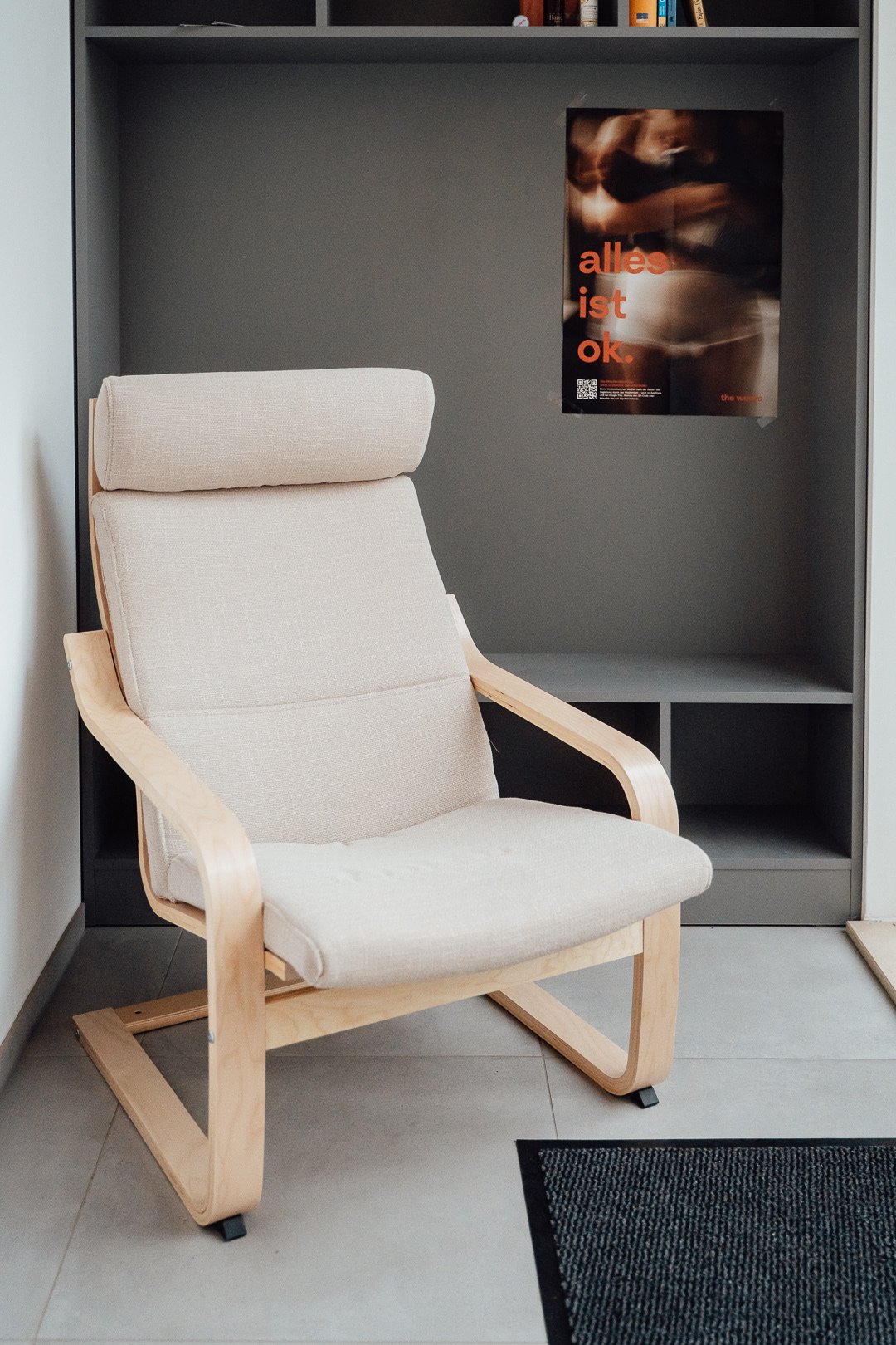 Ein moderner bequemer beige Stuhl mit Holzrahmen steht vor einer grauen Bücherregalwand mit einem Titanic-Poster an der Wand.