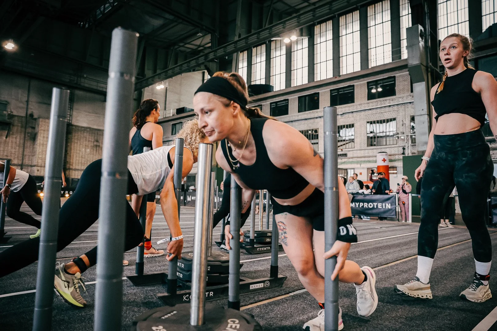 Mehrere Frauen trainieren bei einem Fitness-Event in einer großen Halle, machen Push-Ups unter Hindernissen mit Gewichten.