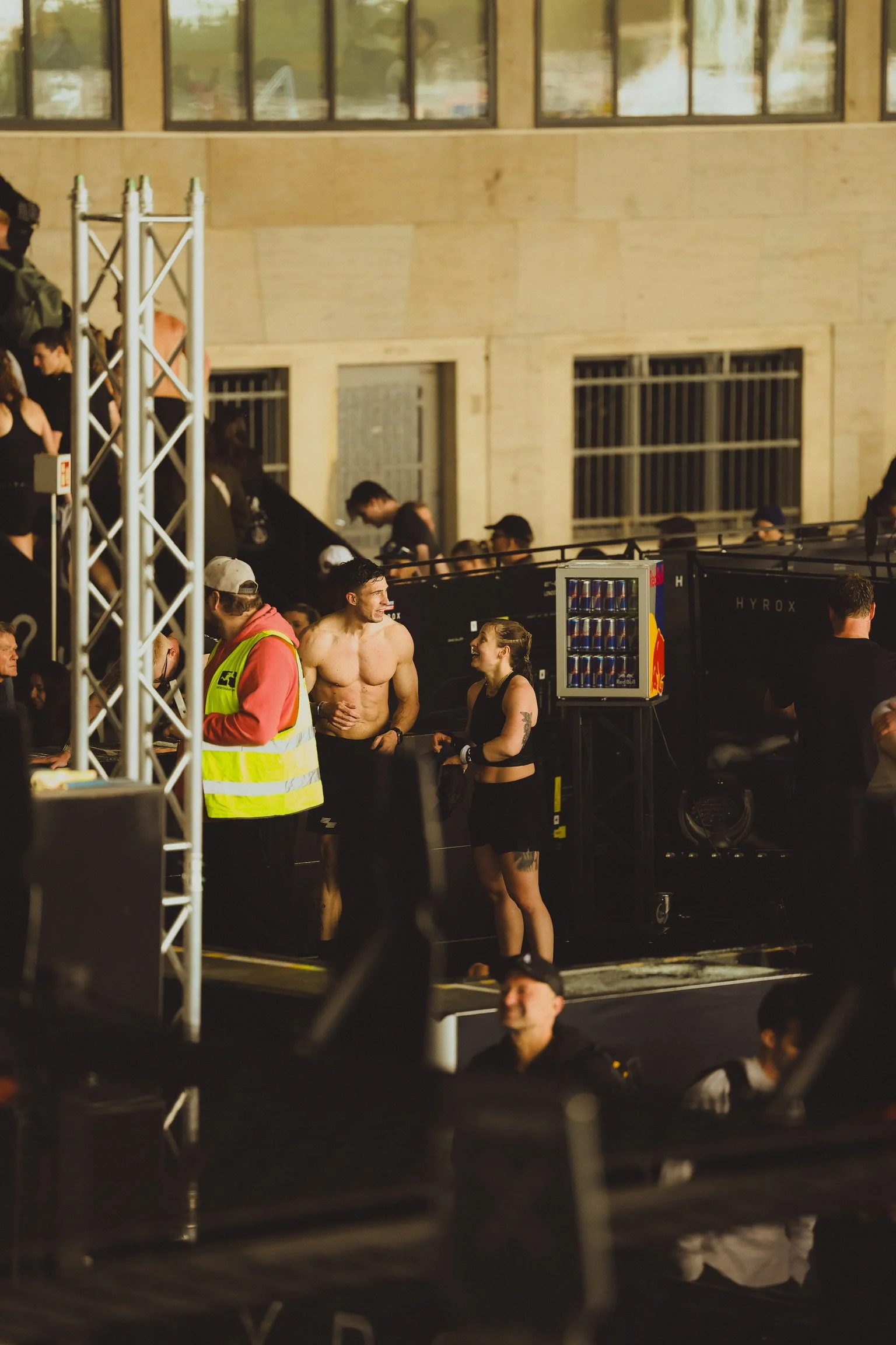 Zwei Menschen mit Sportkleidung und Tattoos, die nach einem Workout oder Event sprechen, während sie vor einer Bühne oder Bühne-Setup stehen. Es gibt mehrere Personen im Hintergrund, einige sind auf den oberen Rängen, und es befinden sich Getränkeautomaten in der Nähe.