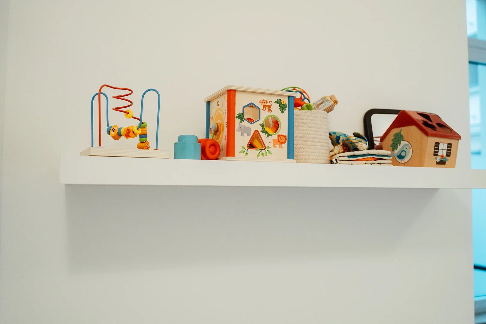 Weißer Wandershelf mit bunten Babyspielzeugen, darunter eine Bead-Table, eine Holzspielhaus-Box, Stoffe und andere Spielsachen.