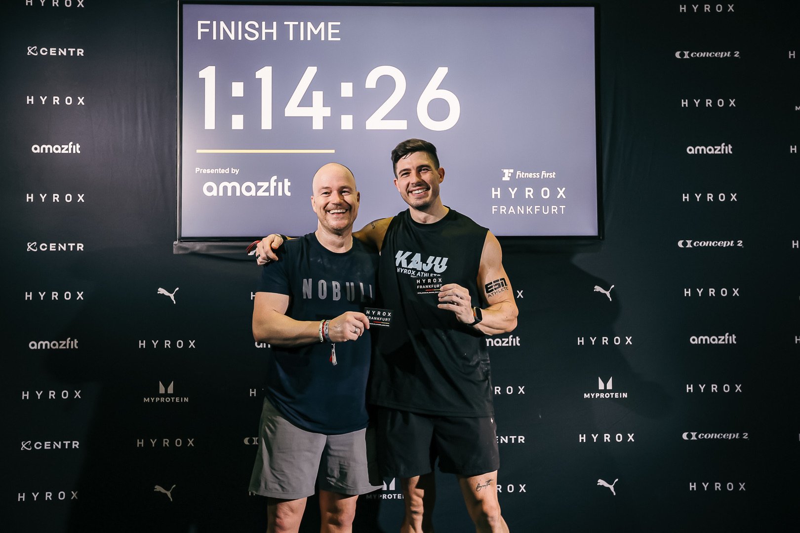 Zwei Männer nach einem Lauf, die lächelnd eine Urkunde und eine Karte halten, vor einem Bildschirm mit der Zeit 1:14:26 bei einem Fitness-Event in Frankfurt.