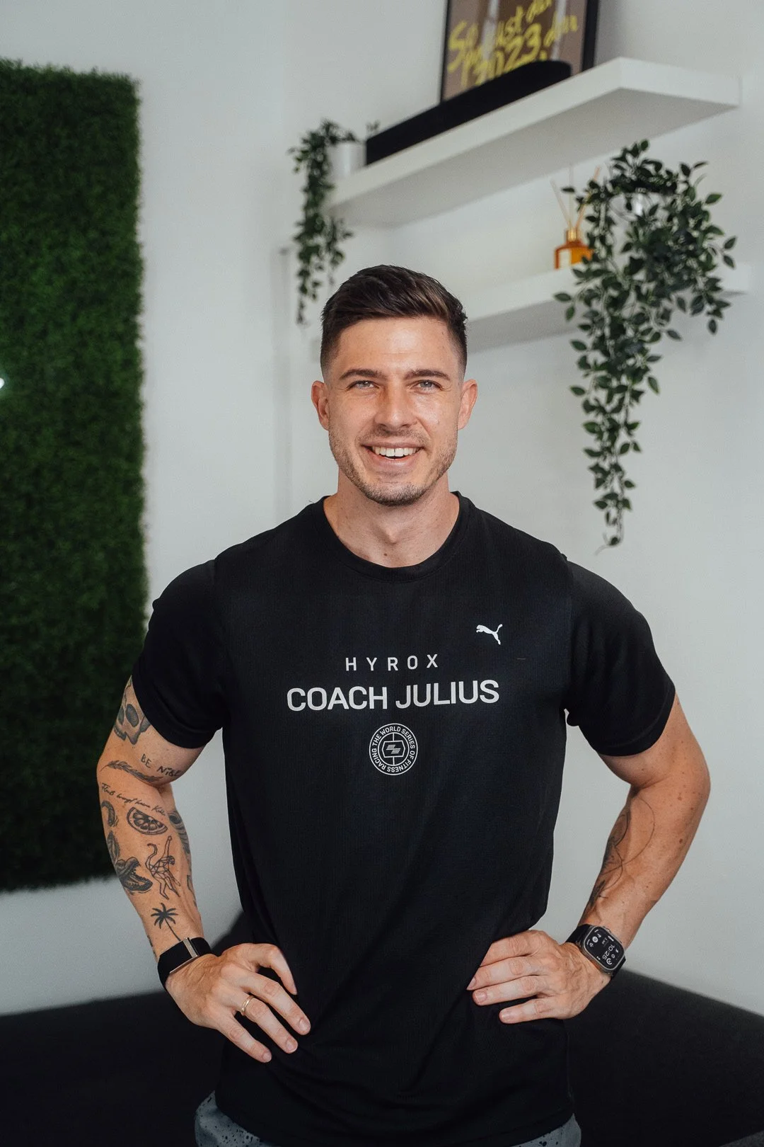 Ein junger Mann steht lächelnd mit Händen in den Hüften in einem Raum mit weißen Wänden, grüner Wandverzierung und hängenden Pflanzen, trägt ein schwarzes T-Shirt mit der Aufschrift "Coach Julius".