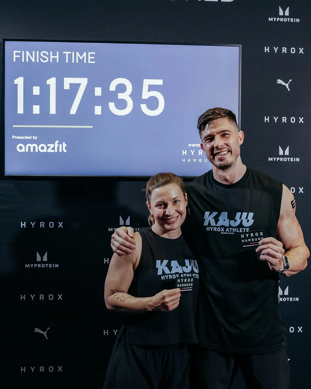 Zwei begeisterte Sportler, eine Frau und einen Mann, nach einem Lauf, tragen schwarze Trikots mit dem Text 'KAU Hyrox Athlete', stehen vor einem schwarzen Hintergrund mit Logos und halten eine Trophäe. Im Hintergrund ein Bildschirm, der die Zeit 1:17:35 zeigt, mit dem Text 'Finish Time' und 'Presented by amazfit'.