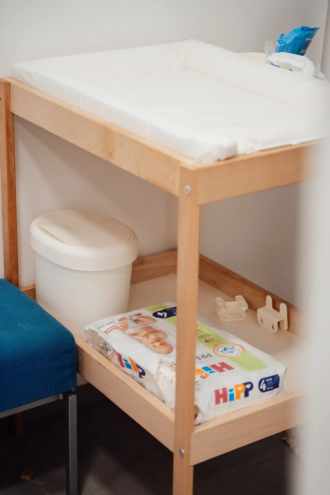 Ein Kinderbett mit Wickelauflage, Wickelauflage, Windel- und Feuchttücherpaket, Mülleimer und Kinderartikel auf einem Holzrahmen, daneben ein blauer Stuhl.