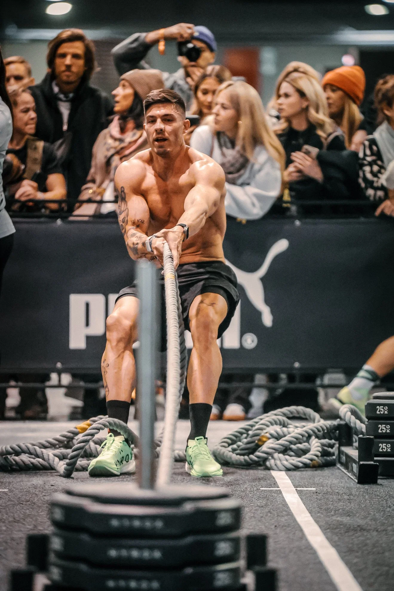 Ein muskulöser Mann beim Google-Workout bei einem Fitnesswettbewerb, umgeben von Zuschauern und einem Fotografen im Hintergrund.