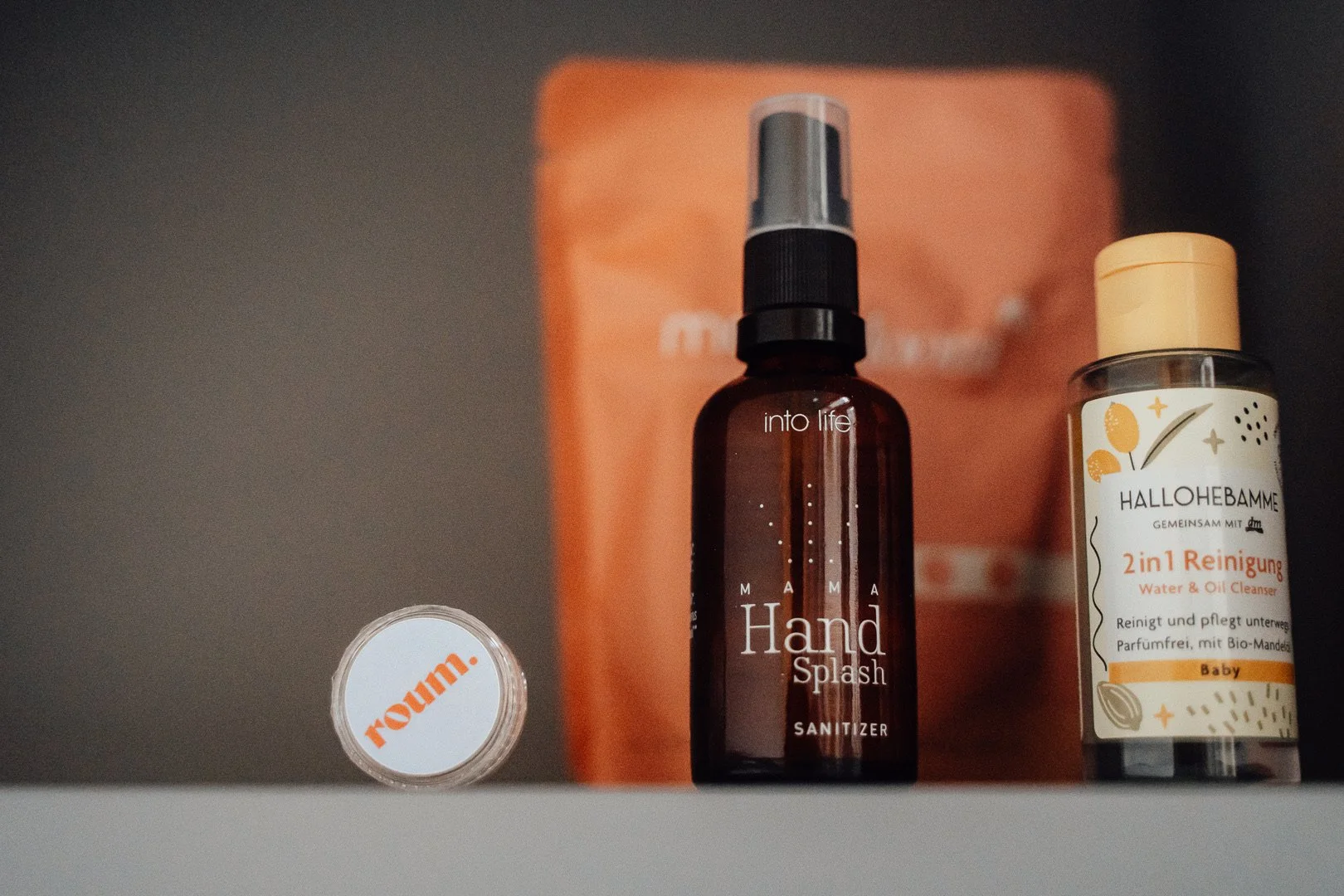 Drei Pflegeprodukte auf einer weißen Oberfläche. Von links nach rechts: Eine kleine Dose mit orangener Aufschrift "Now!" auf dem Deckel, eine braune Sprühflasche mit weißer Beschriftung "Mama Hand Splash" und eine Flasche mit Shampoo "Hallohabamme 2 