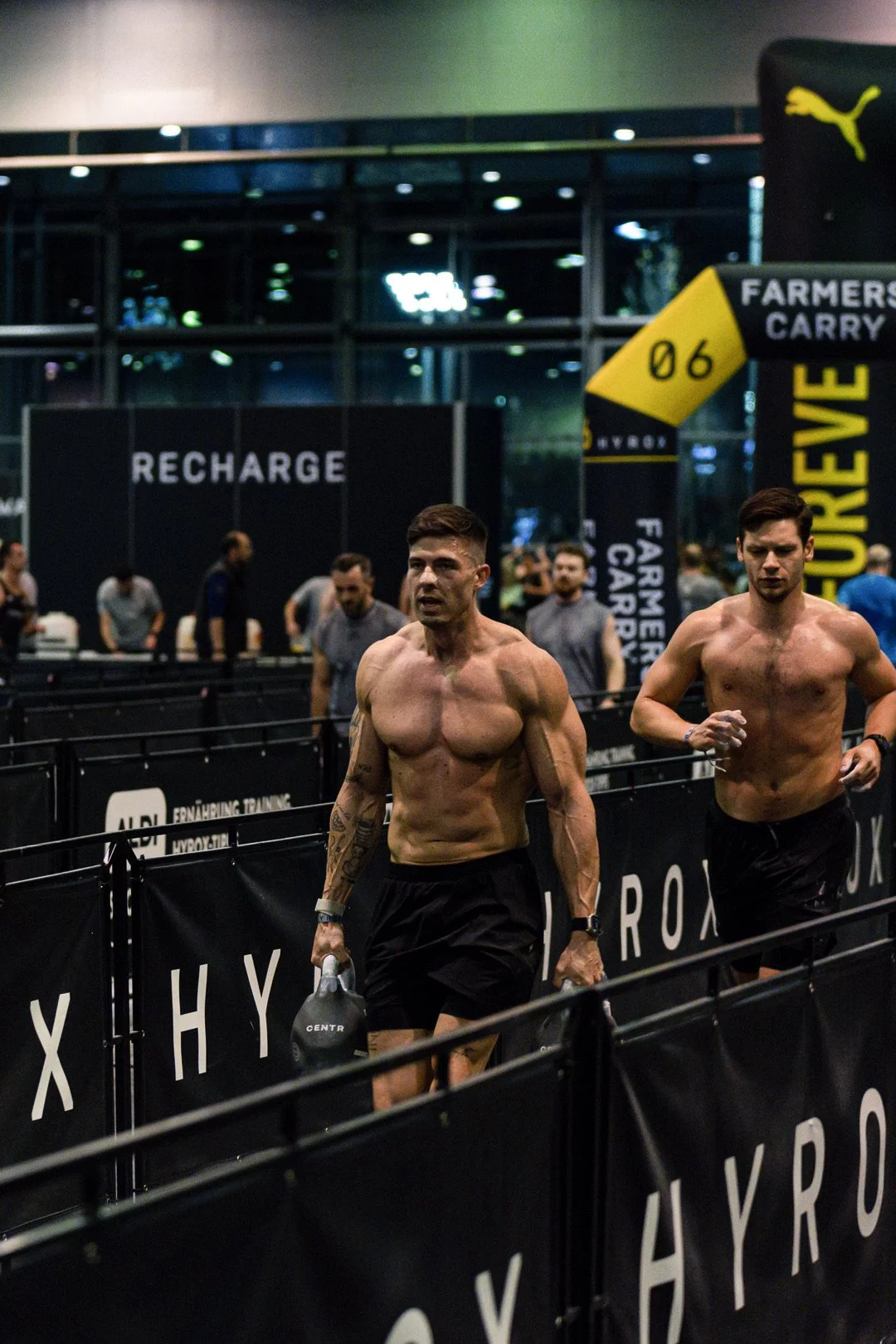 Zwei shirtlose Männer laufen auf einem Laufband in einem Fitnessstudio. Im Hintergrund sind andere Personen und Werbung für das Fitnessstudio zu sehen.