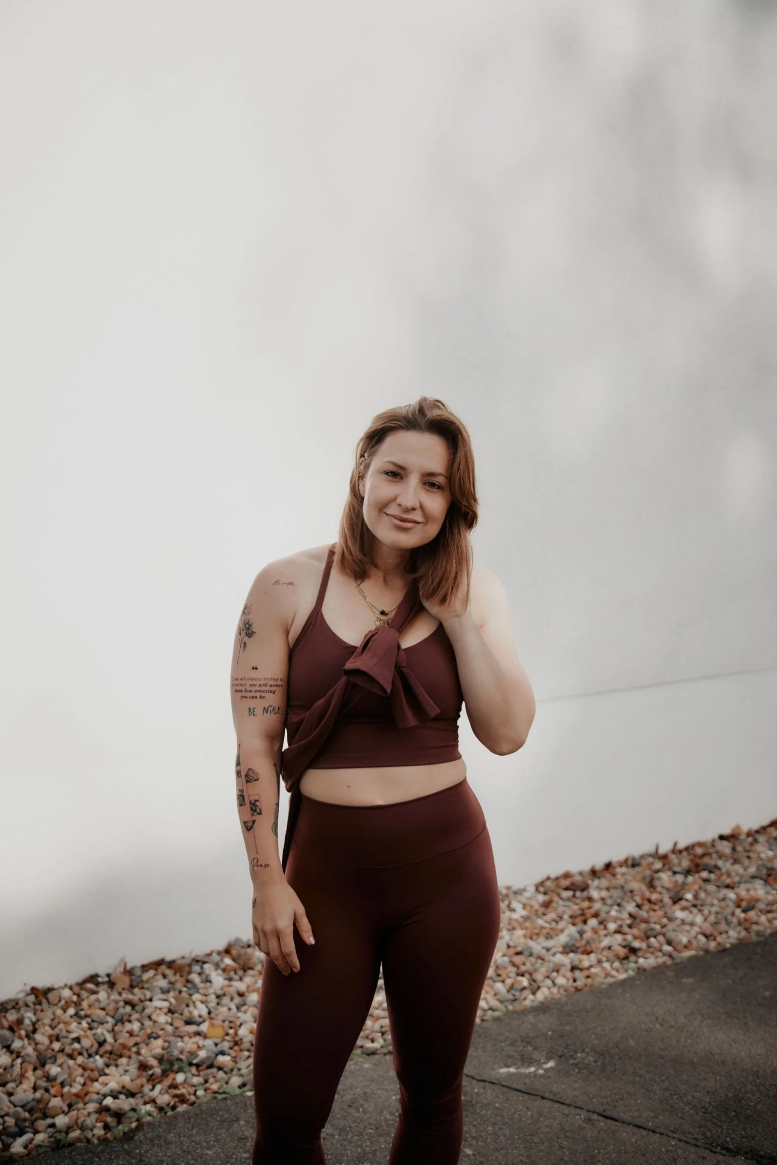 Junge Frau in braunem Sportoutfit steht vor weißer Wand auf Stein- und Asphaltboden, lächelt, hat Tattoos am Arm, trägt Halskette, und hat braune, lockige Haare.