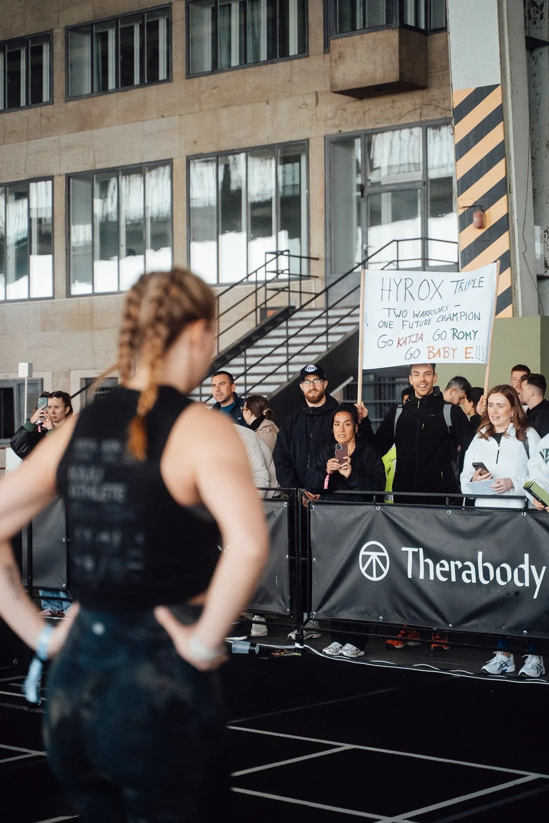 Eine Frau im Vordergrund, die auf einem Sportevent steht, während eine Gruppe von Zuschauern im Hintergrund eine supportive Haltung einnimmt, einige mit Mobiltelefonen in der Hand. Es gibt ein Schild mit der Aufschrift 'HYROX TRIPLE' und eine Anfeuer