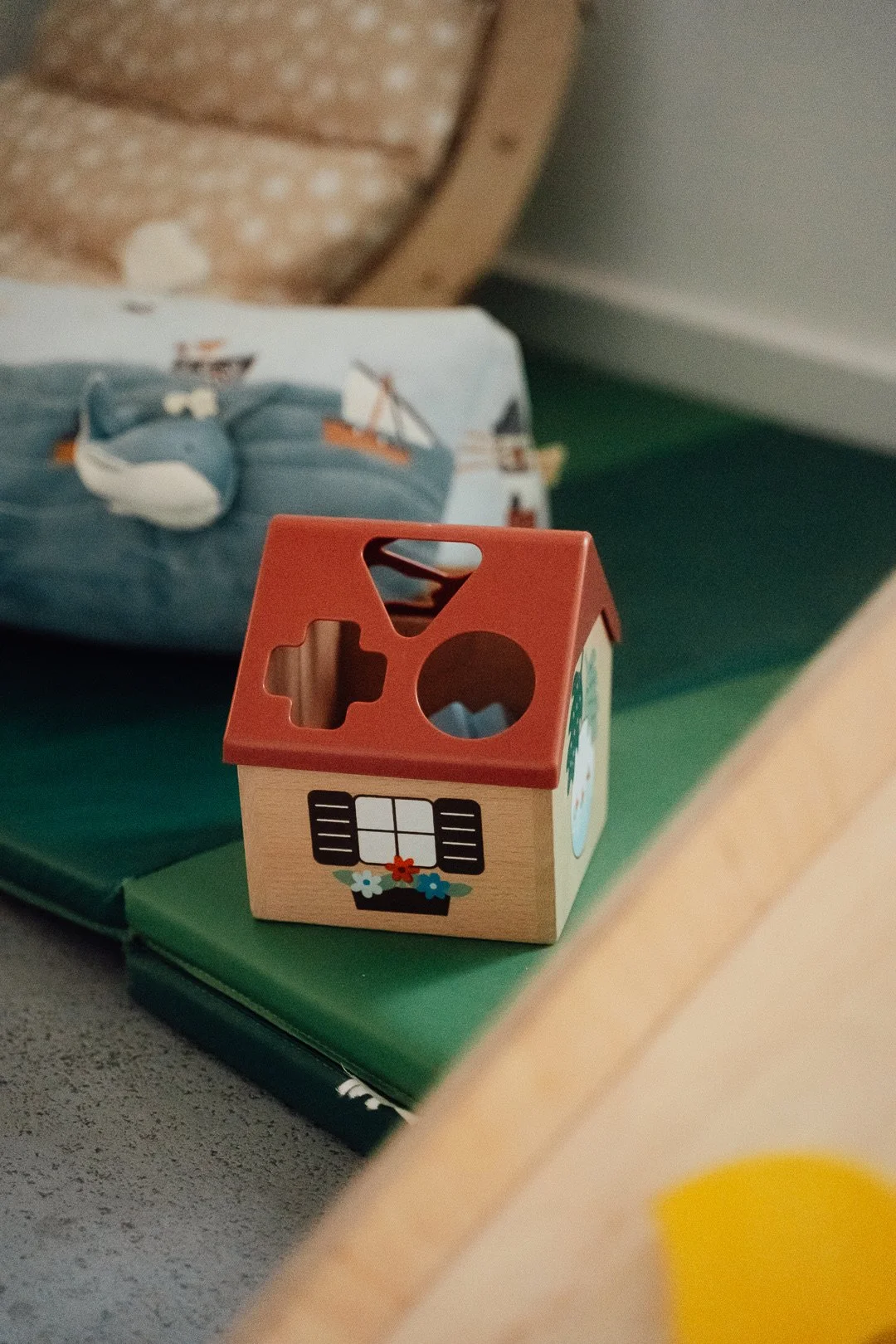 Ein kleines hölzernes Spielzeughaus mit roten Dach und Fensterdekoration steht auf einer grünen Oberfläche, im Hintergrund ist ein Kissen mit Schiffsmotiv zu sehen.