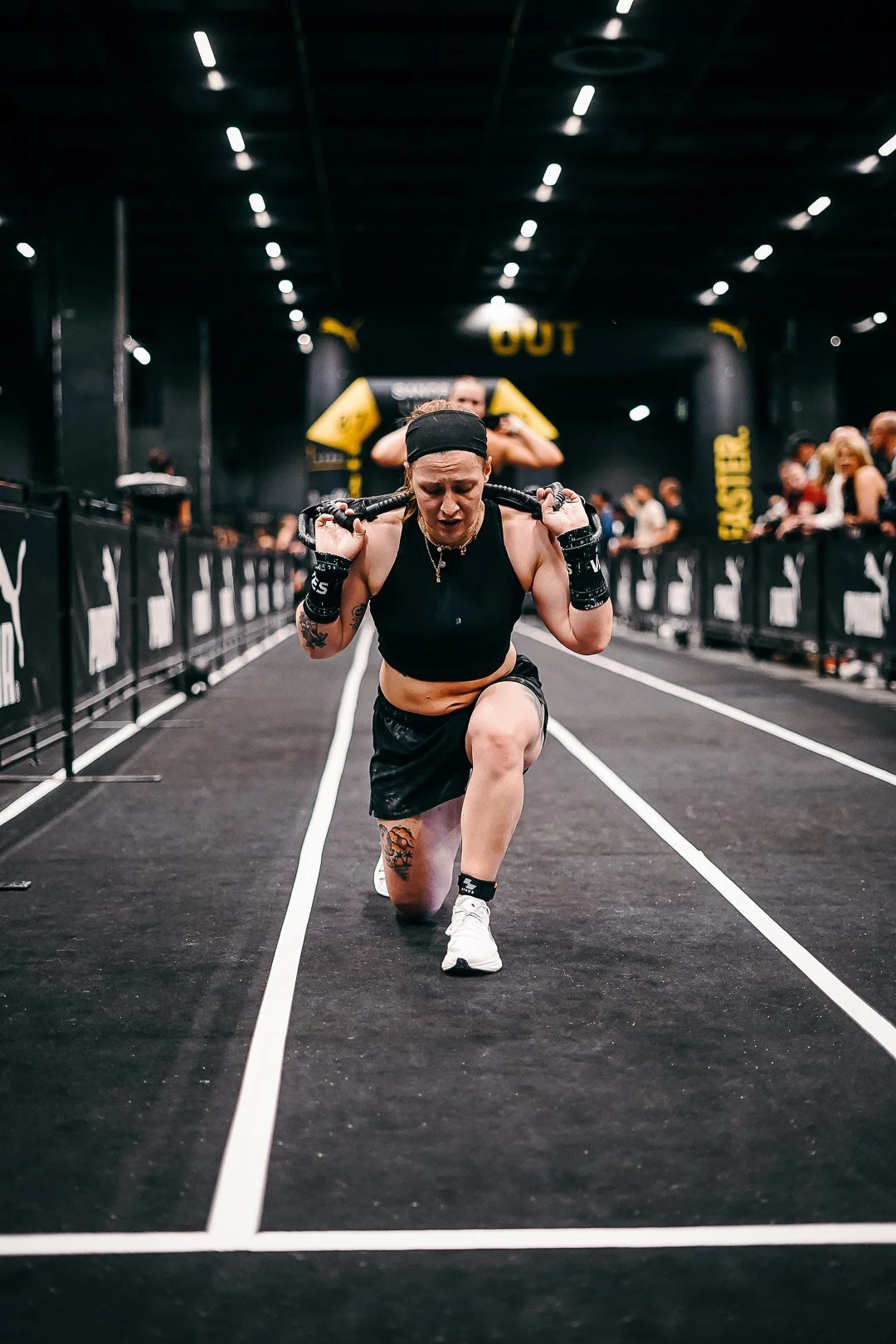 Eine sportliche Frau beim CrossFit-Wettkampf, sie kniet auf einer schwarzen Laufbahn, trägt eine schwarze Sportbekleidung und hebt eine Schlaufe auf ihren Schultern, während sie von Zuschauern im Hintergrund beobachtet wird.