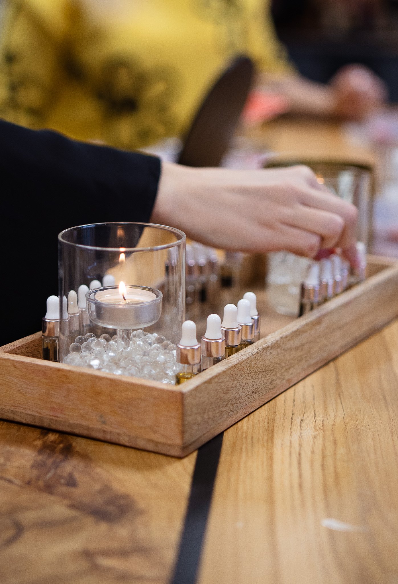 A person's hand reaching for a small bottle on a wooden tray containing multiple small bottles with droppers, a lit candle, and decorative glass beads.