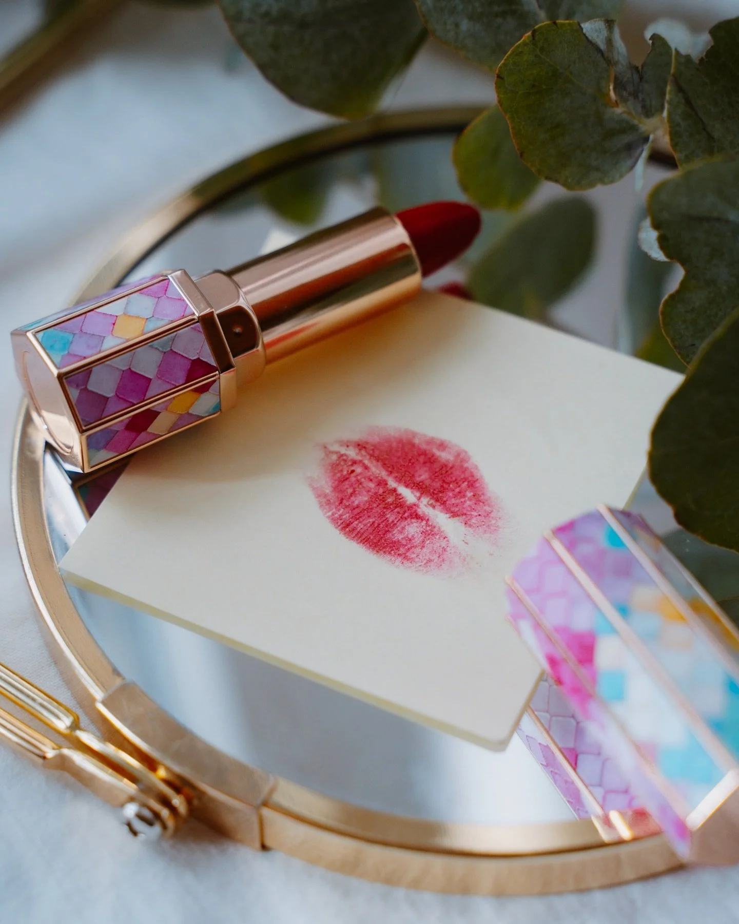 A lipstick with a colorful, mermaid-scale pattern on its case, a note with a red lipstick kiss mark, and green leaves are arranged on a round mirror.