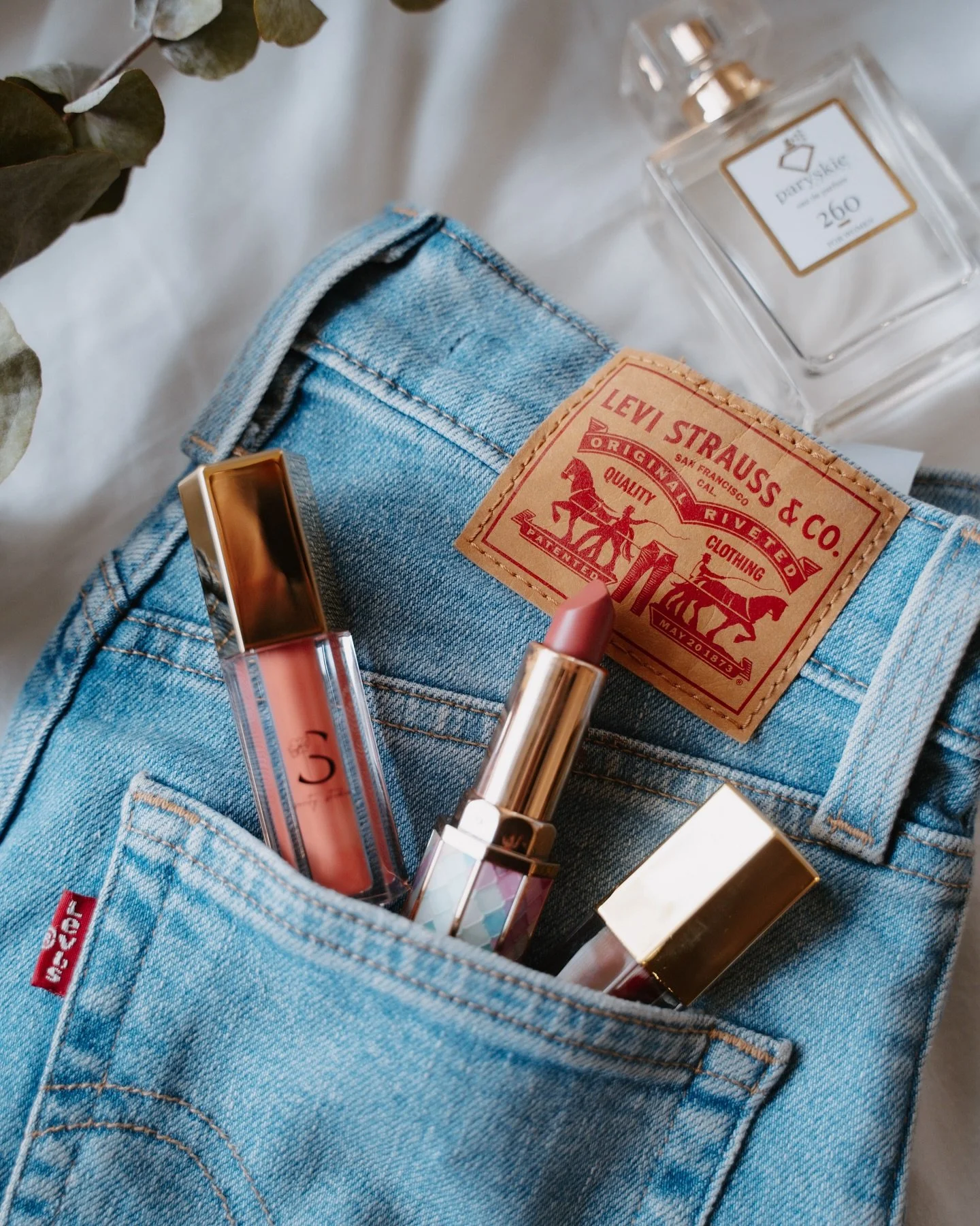 Light blue Levi's denim jeans with a leather patch on the waistband, three makeup lipsticks, and a clear glass bottle of perfume on a white surface with some dried leaves nearby.