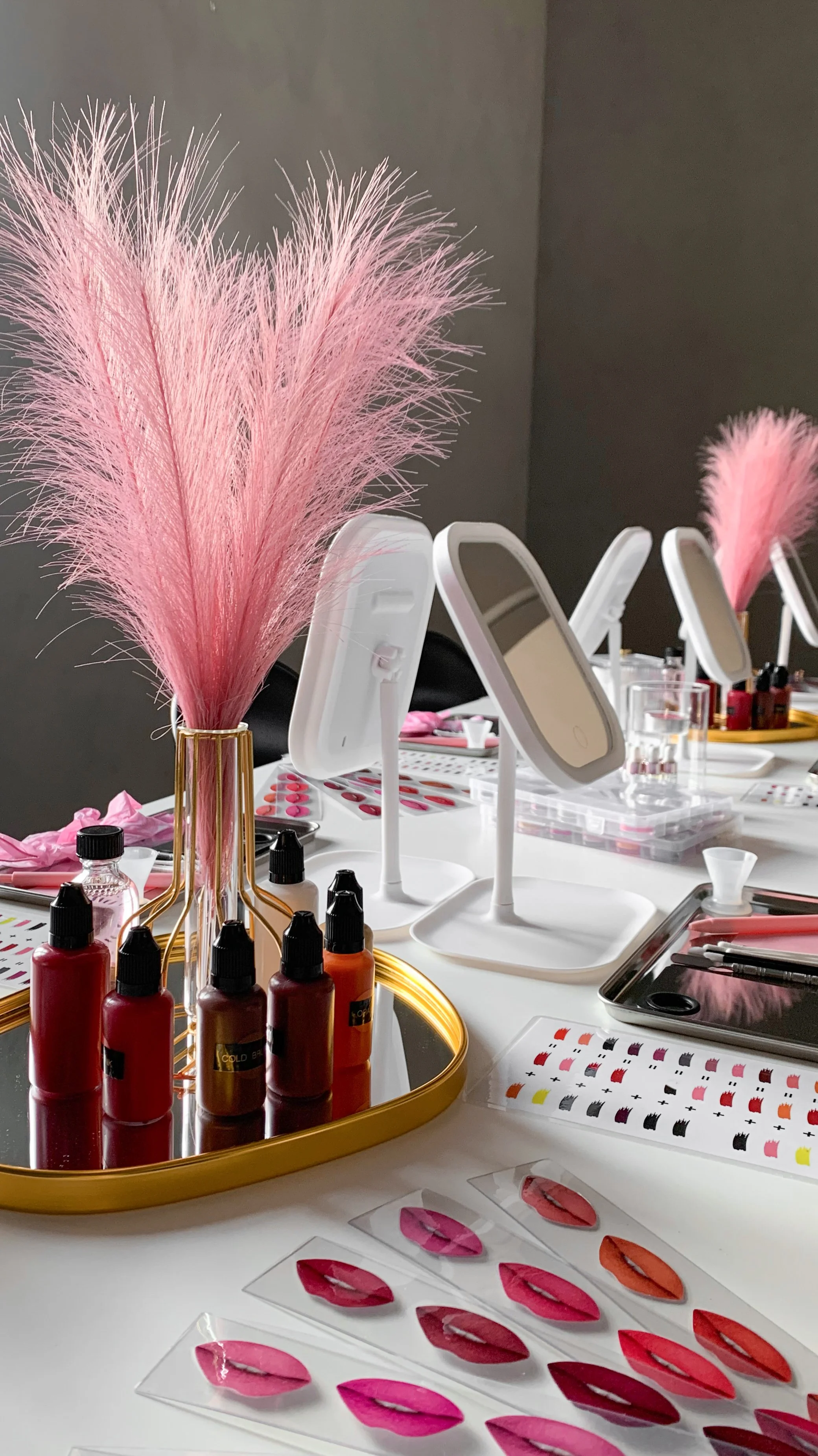 A makeup setup featuring pink fluffy pampas grass in gold vases, multiple mirrors, nail polish bottles, and nail art samples on a white table.