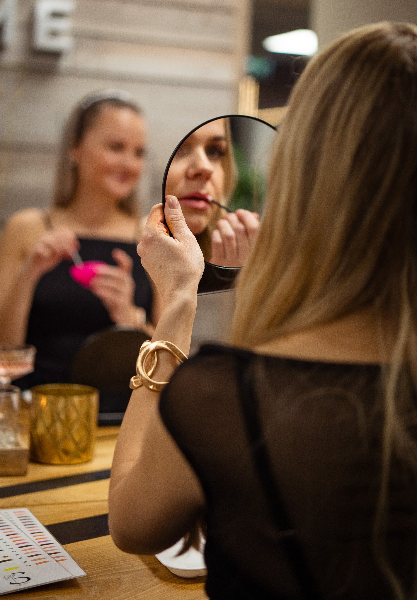 A woman applies lipstick while looking into a small round mirror. She wears a black sheer top and gold jewelry. In the background, another woman holds a pink makeup container and a makeup brush, smiling.