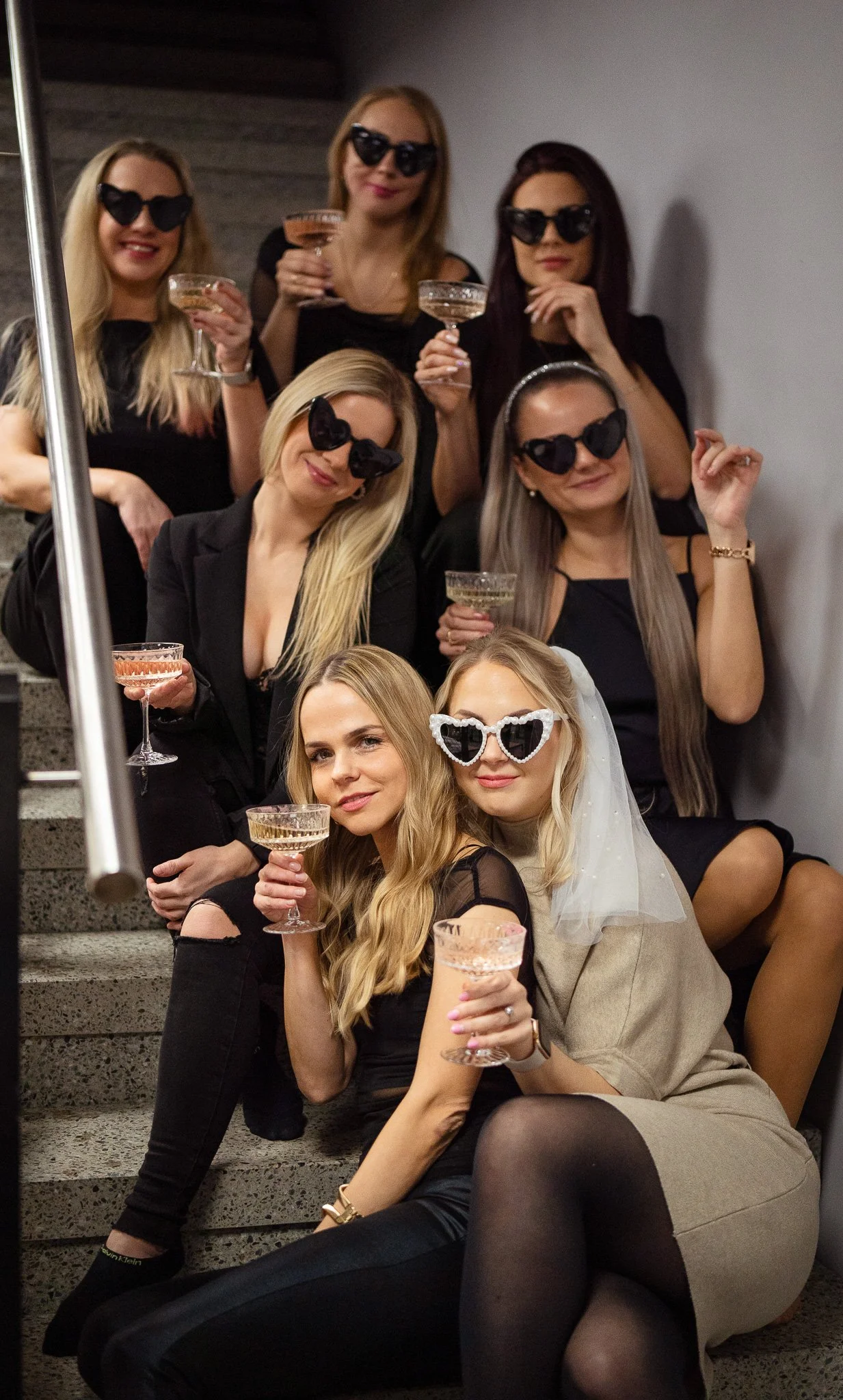 Group of women celebrating with cocktails, wearing sunglasses, some with party accessories, sitting on stairs in a casual party setting.