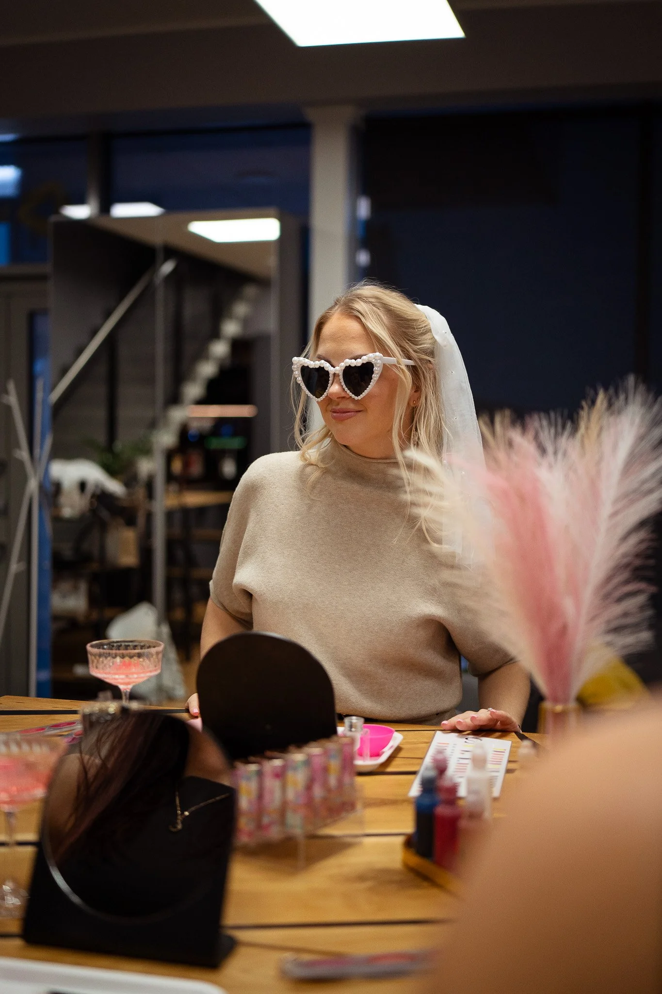A woman with blonde hair wearing white, heart-shaped sunglasses and a beige top, sitting at a table with pink decorations and pink dessert glasses in a modern indoor space.