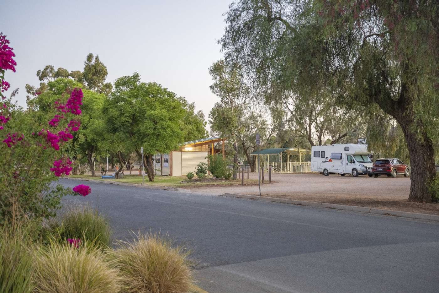 A quiet campground with trees, a camper, a car, and a small building, all under a clear sky.