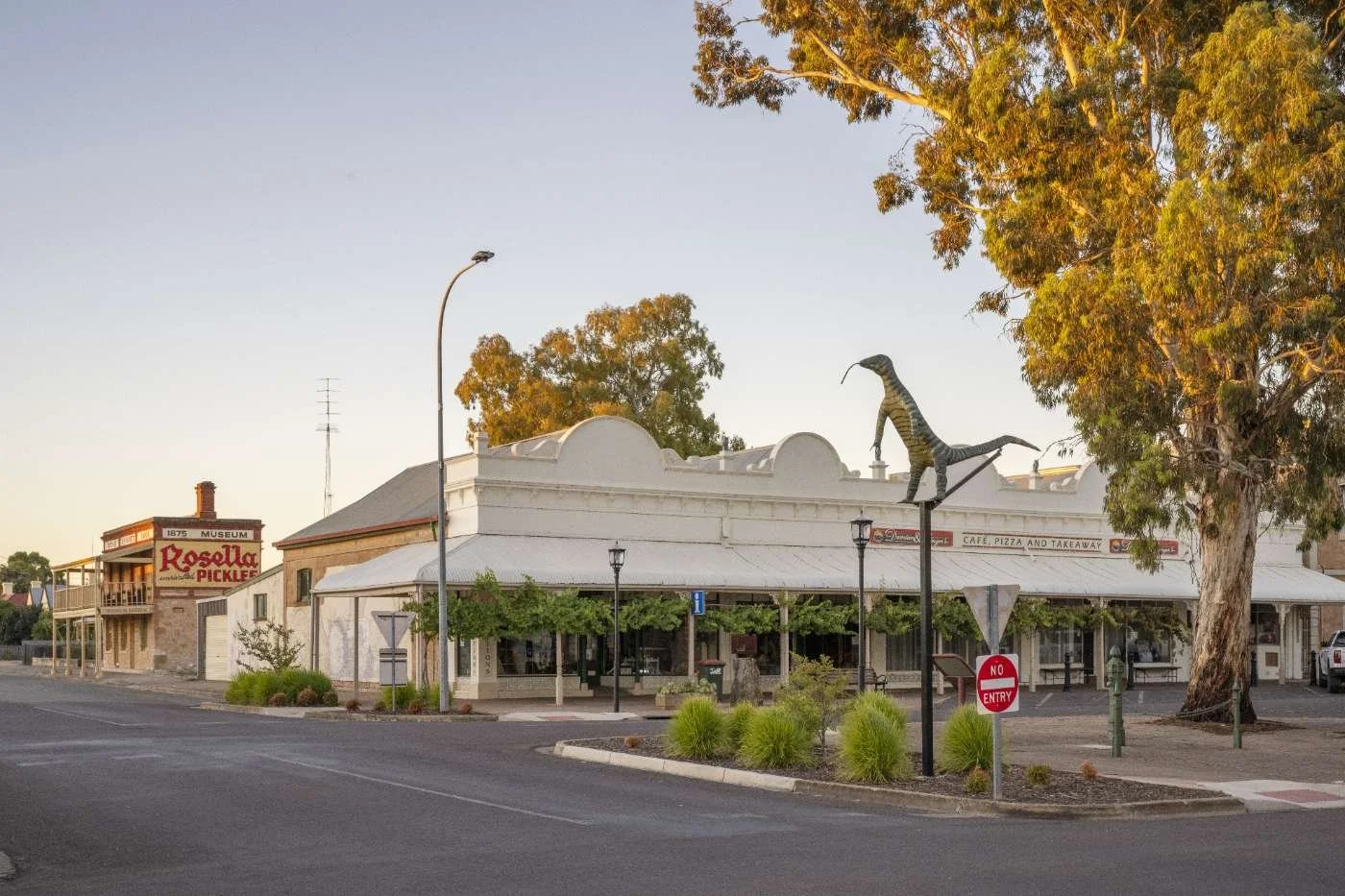 A small town corner with a white building featuring a restaurant sign, trees, two dinosaur sculptures mounted on poles, and a street with a 