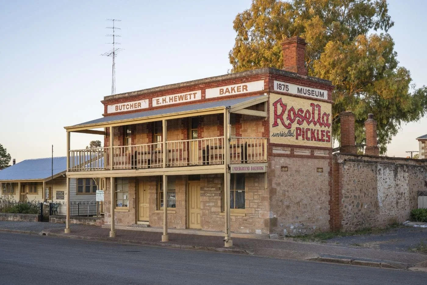 Historic two-story brick building with a balcony, sign reading 'Rosella Pickles' and advertisements for butcher, baker, and museum. Tree in background, street in front.