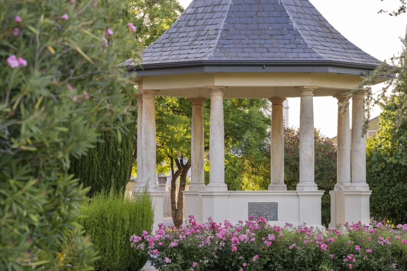 Stone gazebo with columns, surrounded by lush greenery and pink flowers, in a park or garden setting.