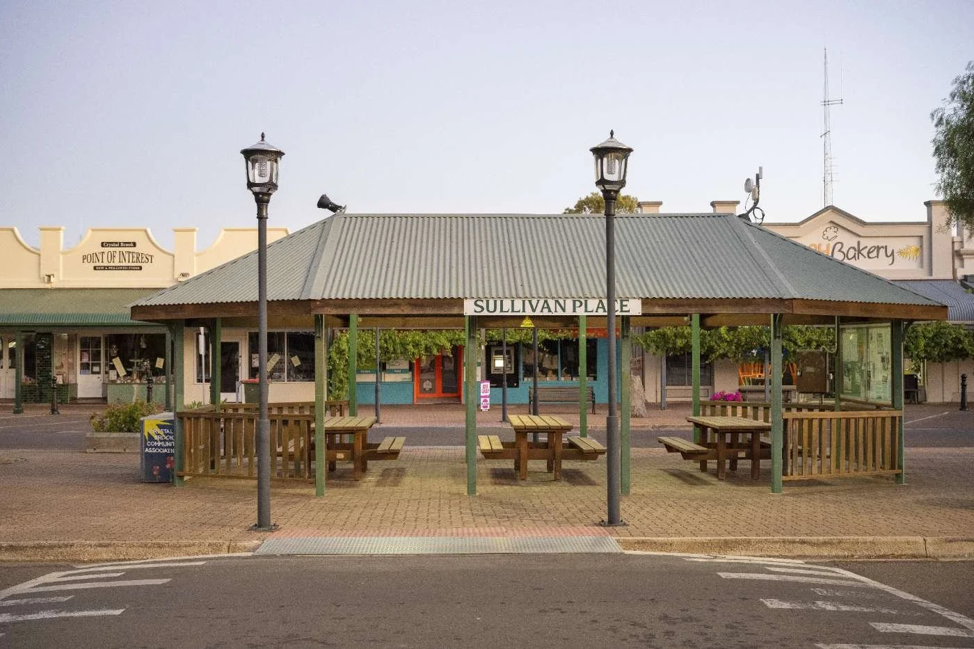 Public pavilion shelter with wooden benches and tables on Sullivan Place, vacant street view in a small town with shops and lampposts.