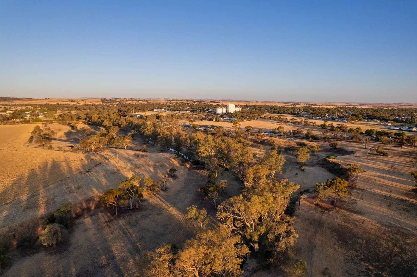 Aerial view of a rural farmland landscape with trees, fields, and distant farm buildings under a clear sky at sunset.