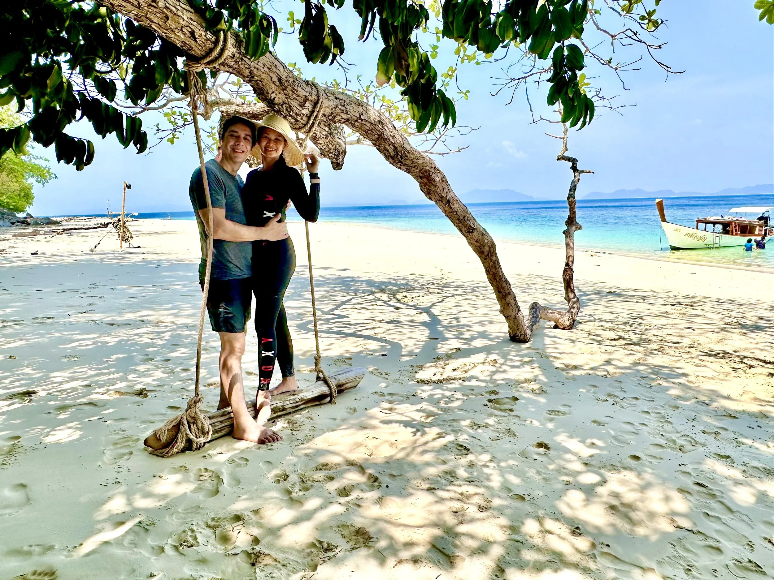 A couple standing on a wooden swing hanging from a tree on a sandy beach, with the ocean and boats in the background on a sunny day.