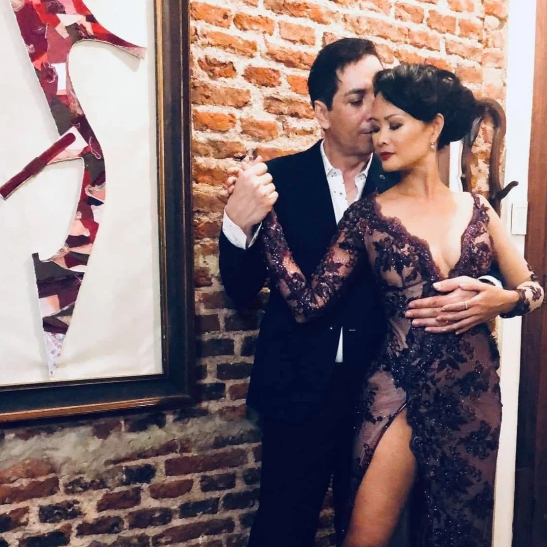 A man and woman dancing intimately, dressed in formal attire, with a brick wall background and framed artwork nearby.