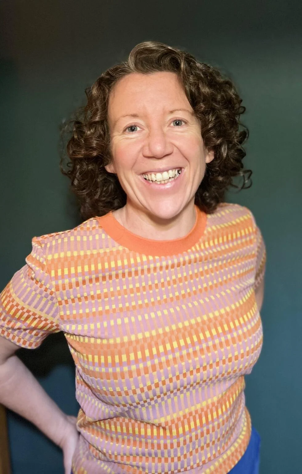 White woman with dark brown curly hair standing and smiling at the camera