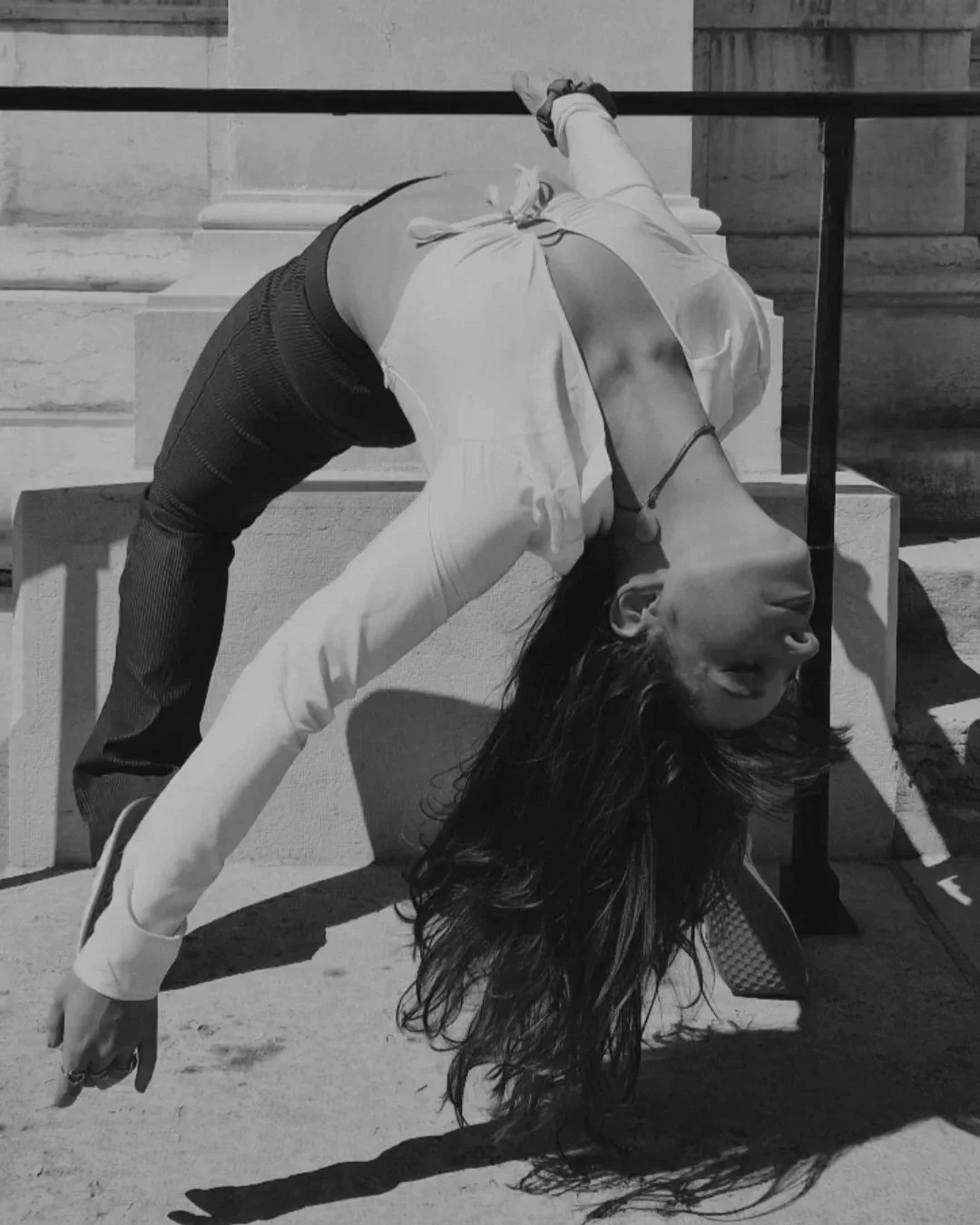 Une femme effectuant une pose de yoga ou de danse, penchée en arrière avec ses cheveux longs pendants vers le sol, en extérieur près d'escaliers en pierre.