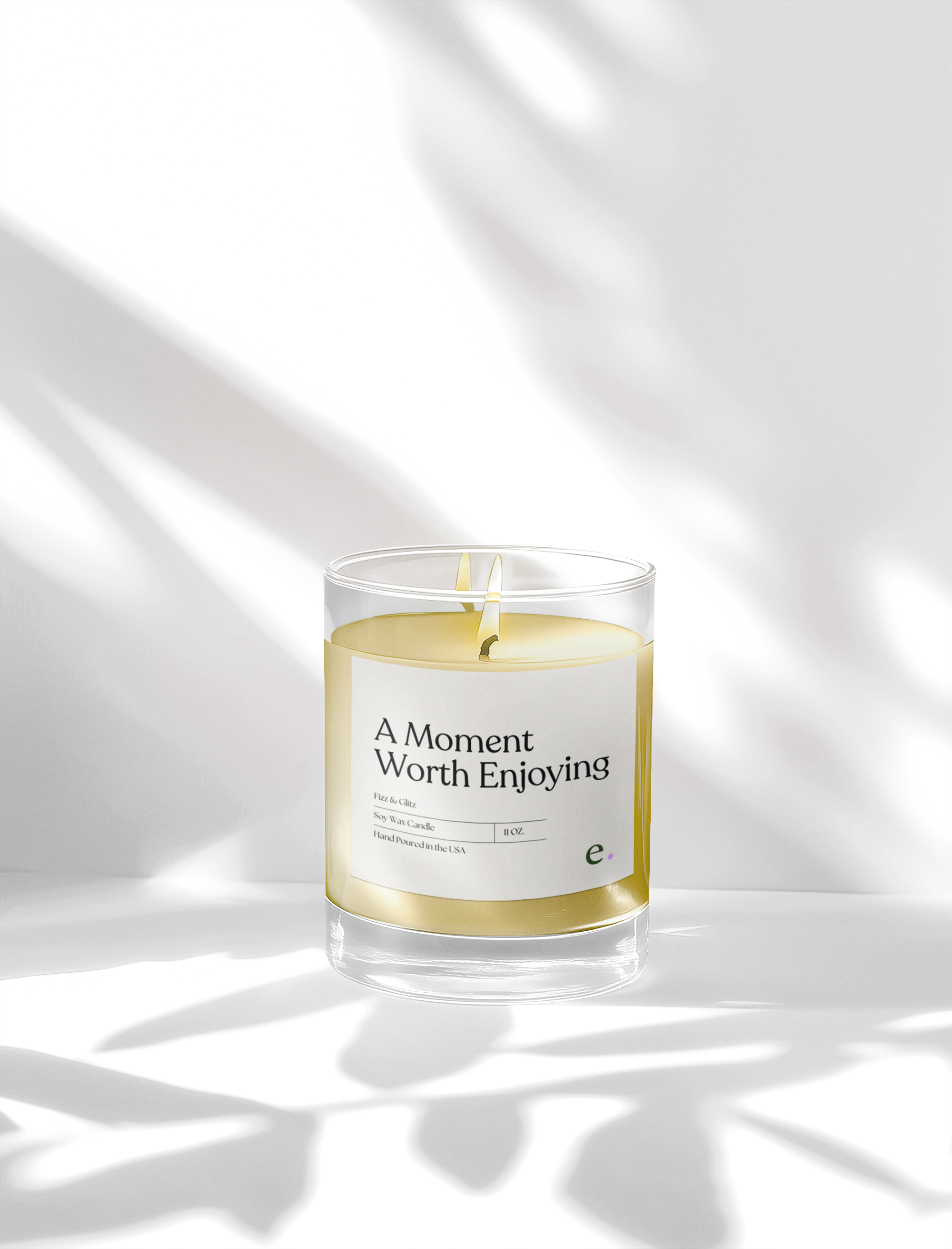 Clear frosted glass candle labeled “A Moment Worth Enjoying,” featuring white wax and photographed in soft sunlight on a clean white background.