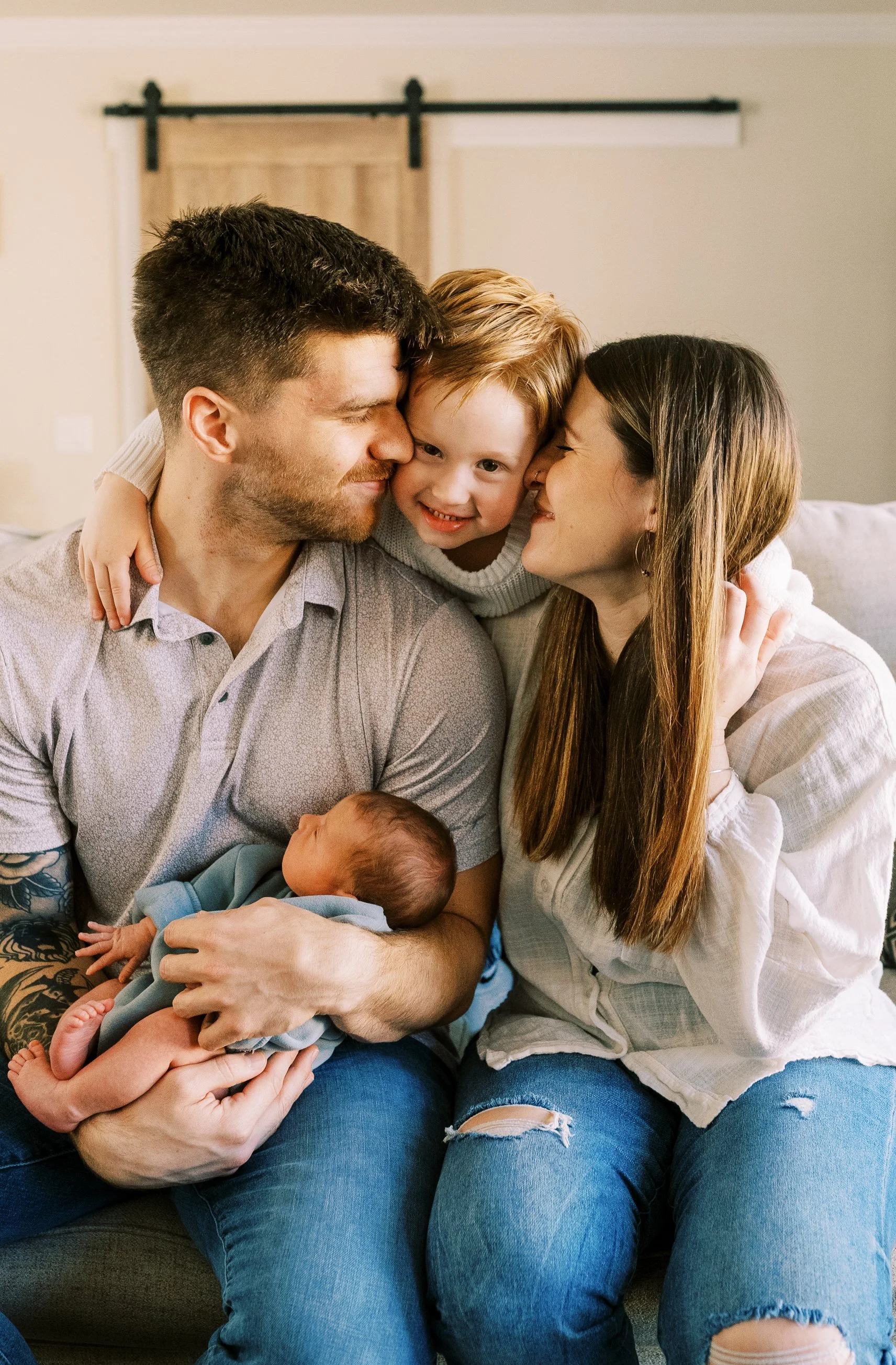 A family of four sitting together on a couch, showing affection. The father is holding a newborn, the mother and a young son are leaning in close, and they are all smiling and touching noses.