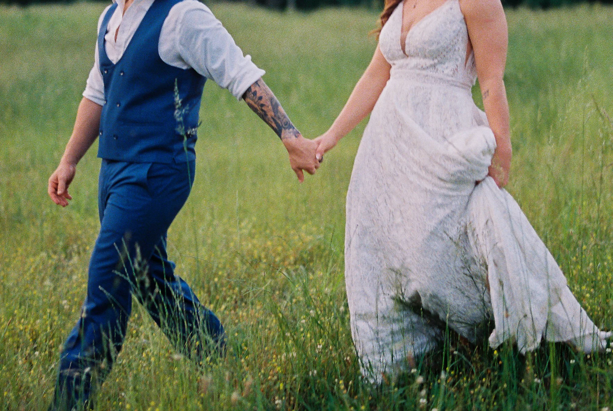 Close crop of the bride and groom walking hand in hand through tall grass, bride lifting the hem of her dress