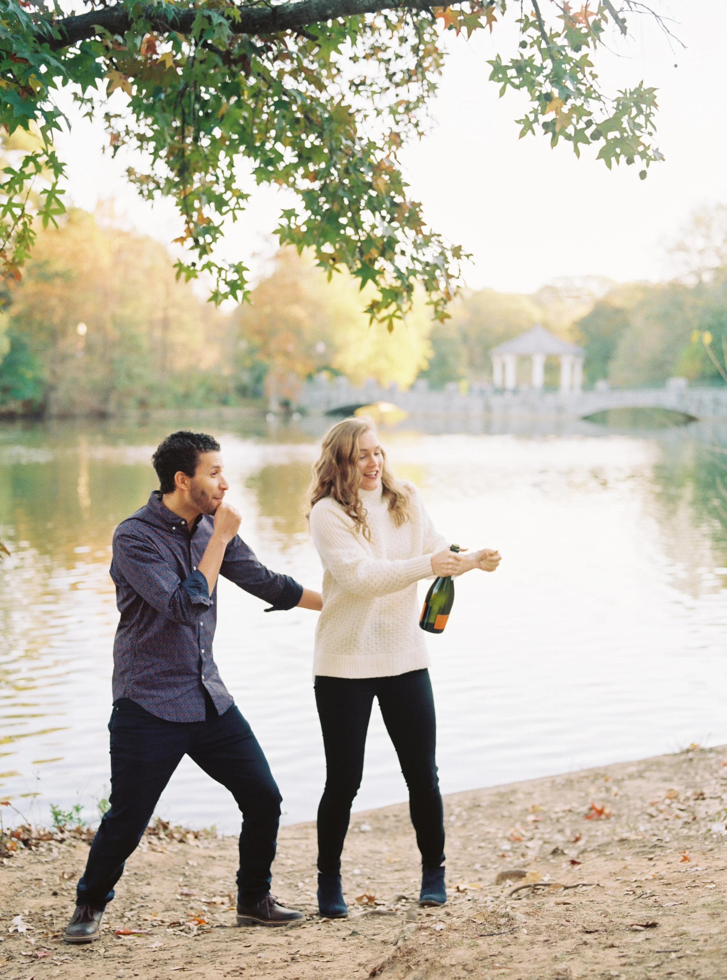 Playful moment of the couple popping a champagne bottle by the lakeshore