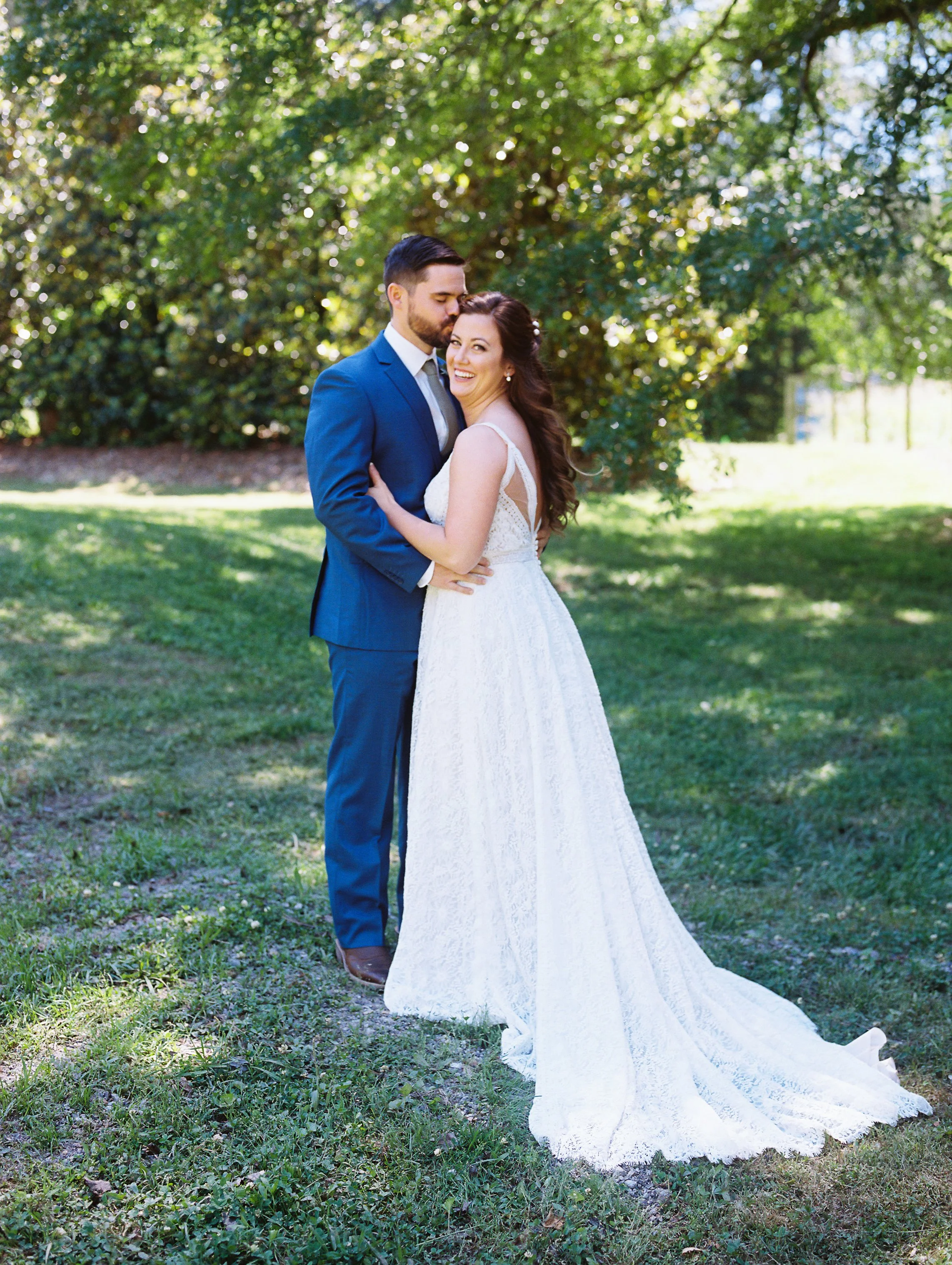 Full-length portrait of the bride and groom standing together under a shaded tree, her gown train spread on the grass