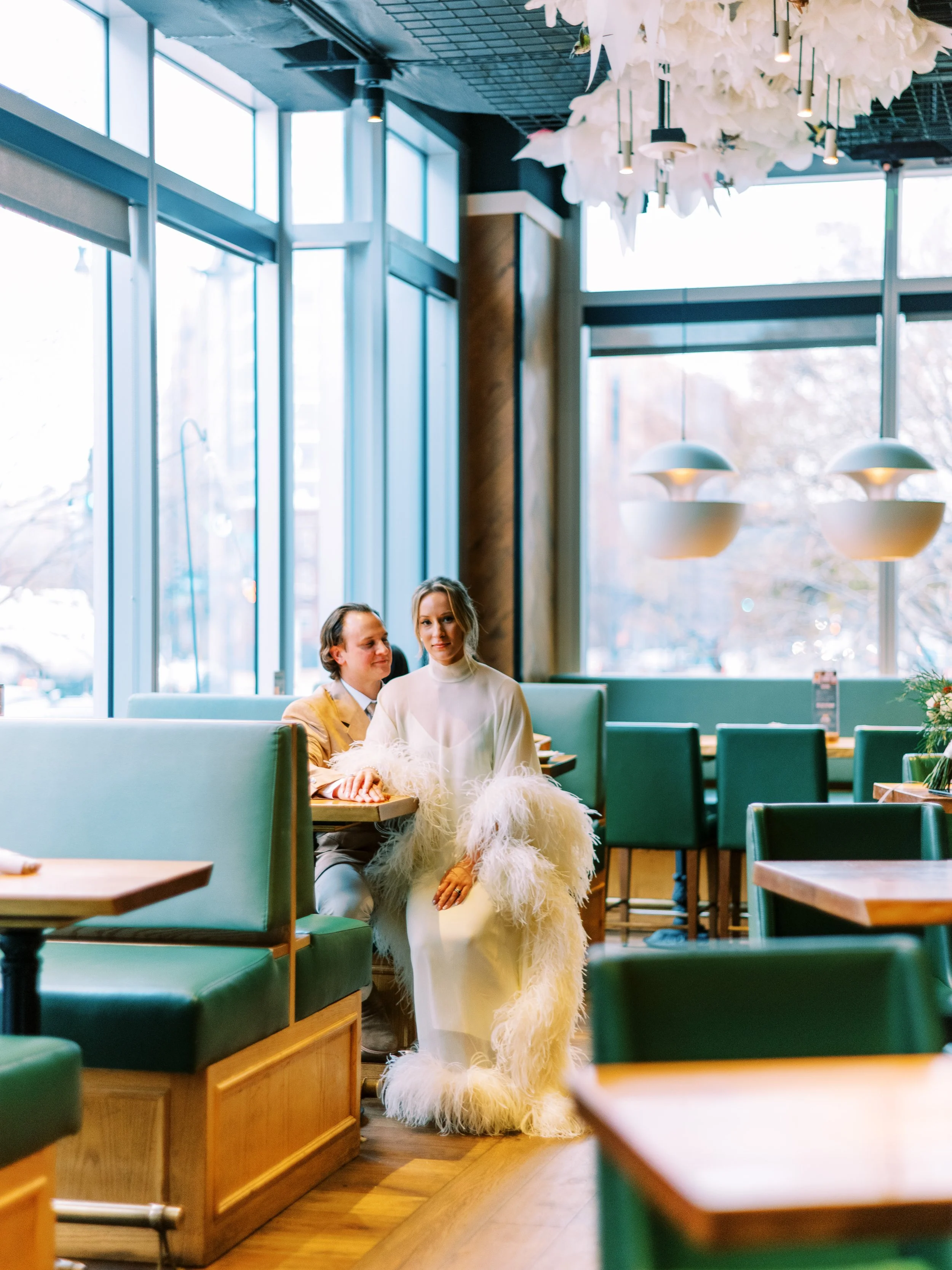 A woman in a white dress with feathered sleeves sitting in a green booth at a restaurant, with a man in a yellow jacket sitting beside her, both by a large window with natural light.