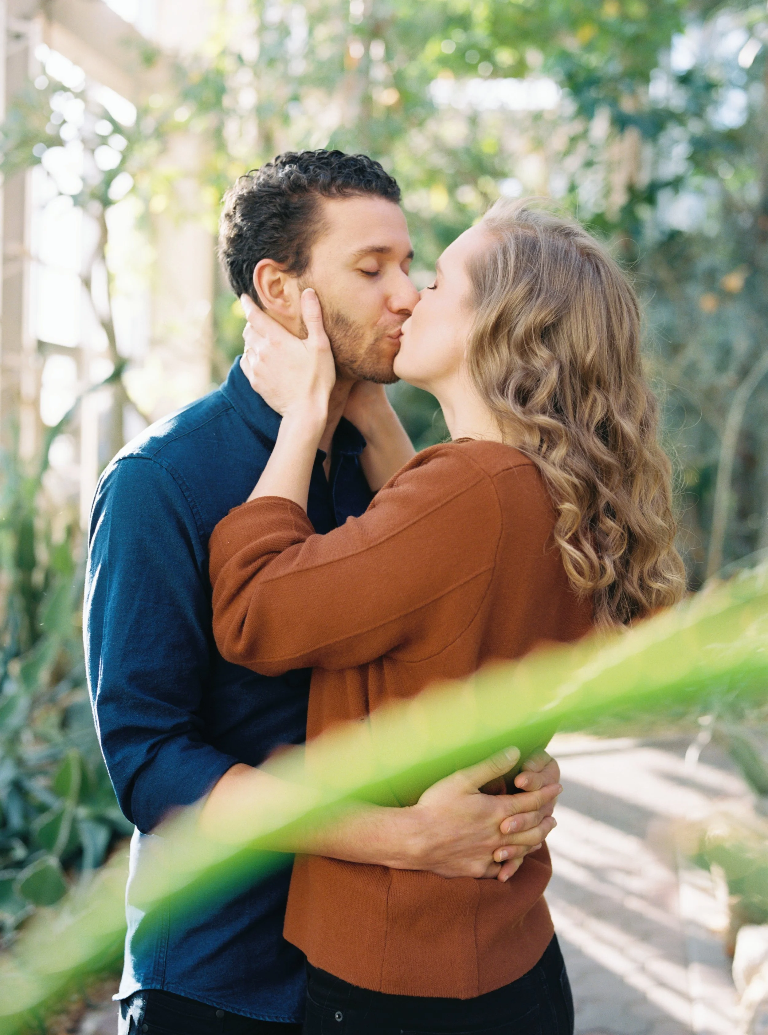 Couple sharing a kiss on a bridge, framed by a green leaf in the foreground