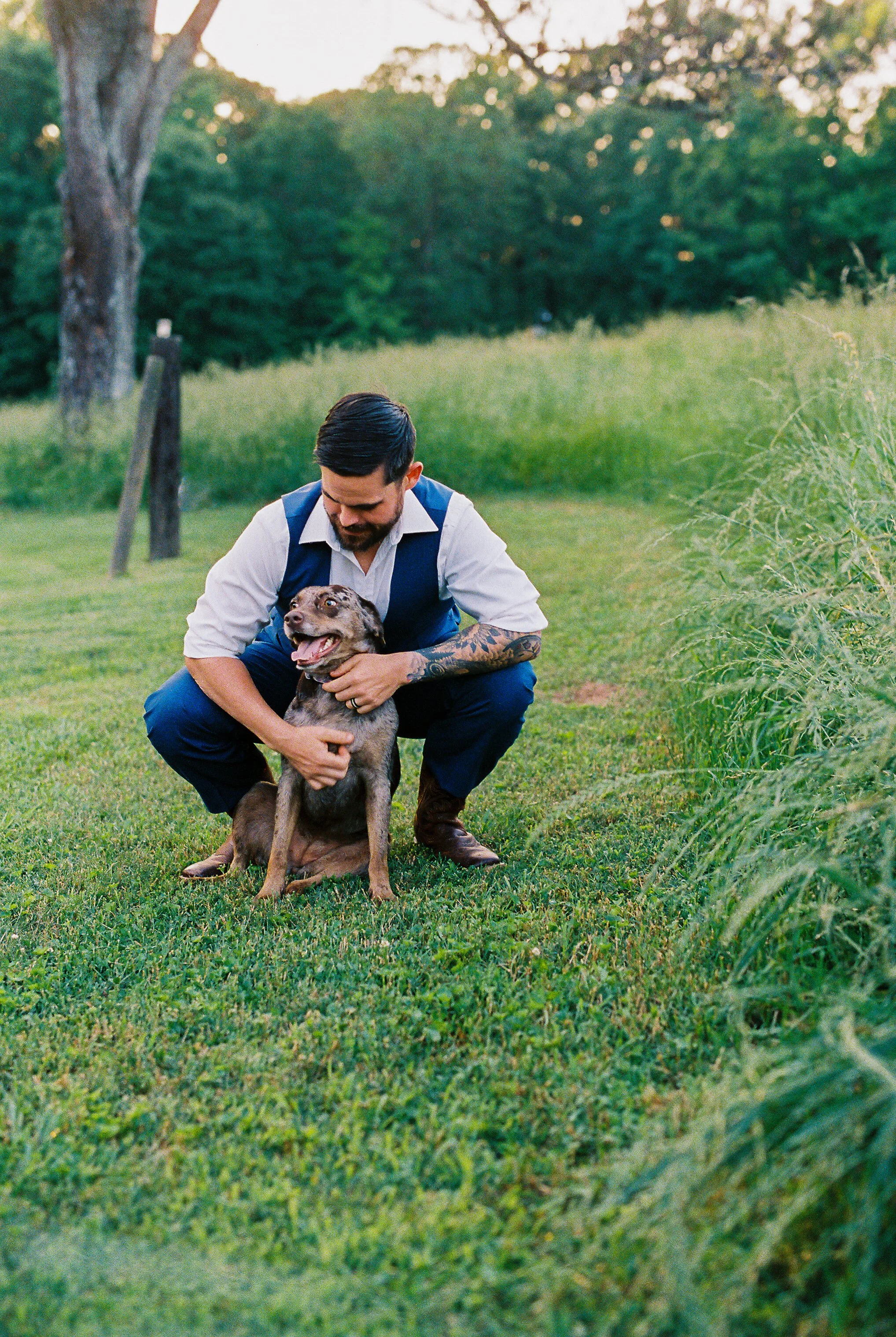 Groom kneeling in a grassy field affectionately petting Dakota the dog