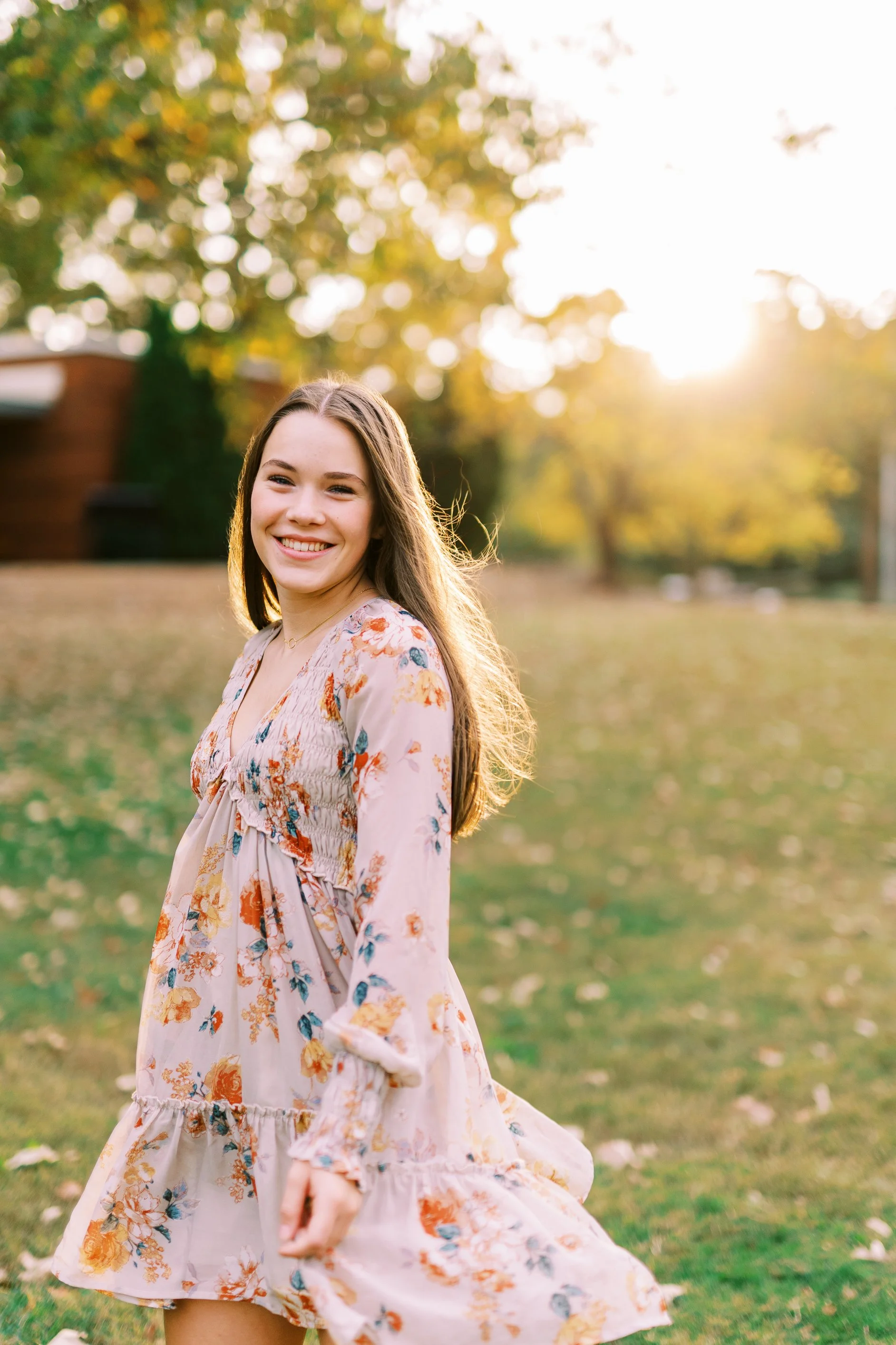 Senior portrait of Hannah glancing back in a floral dress with sunlit trees behind her at Kennesaw Mountain Battlefield Park in Kennesaw, Georgia