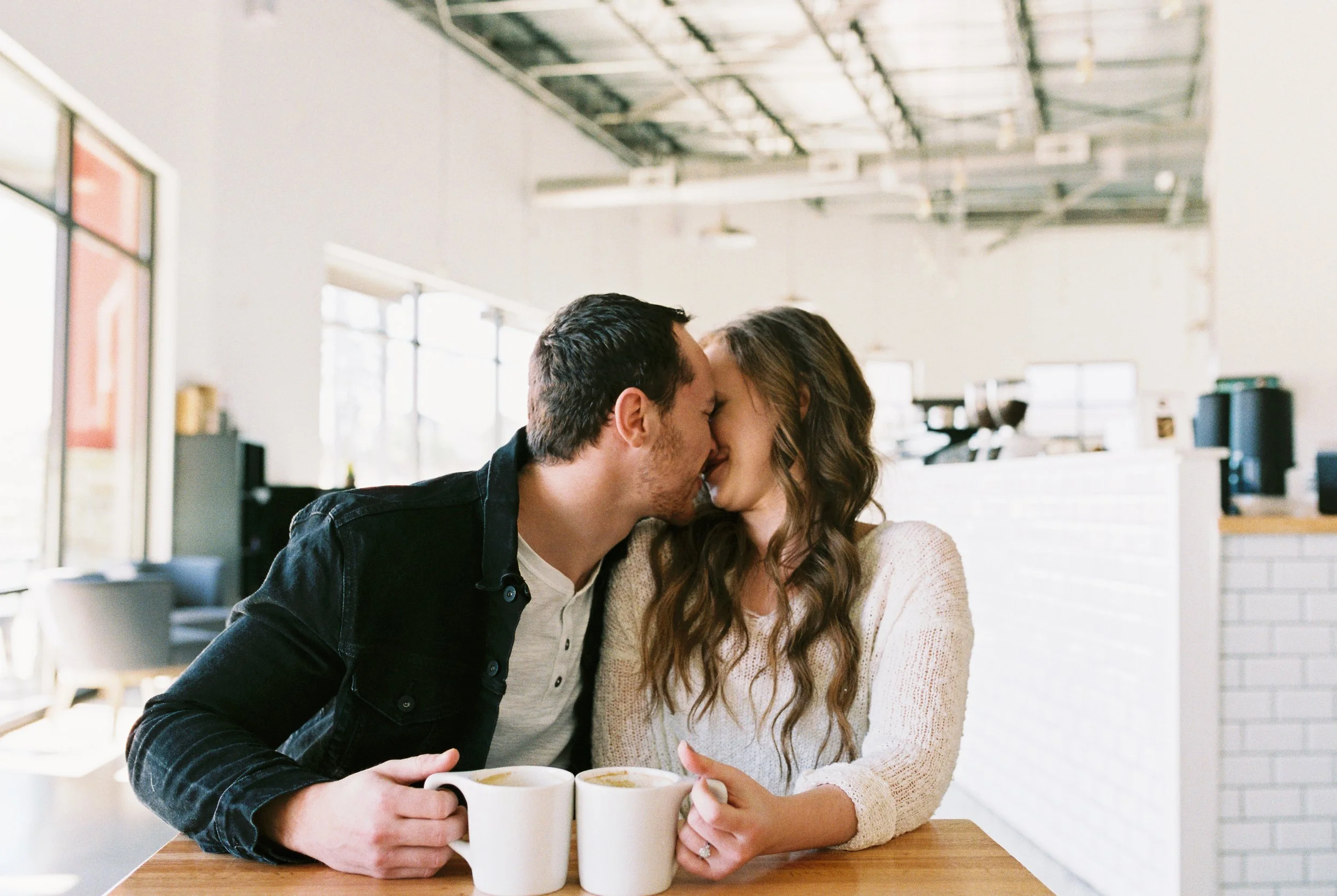 Conner and Maddie kiss over coffee mugs at a wooden table in a Dallas, Georgia coffee shop.