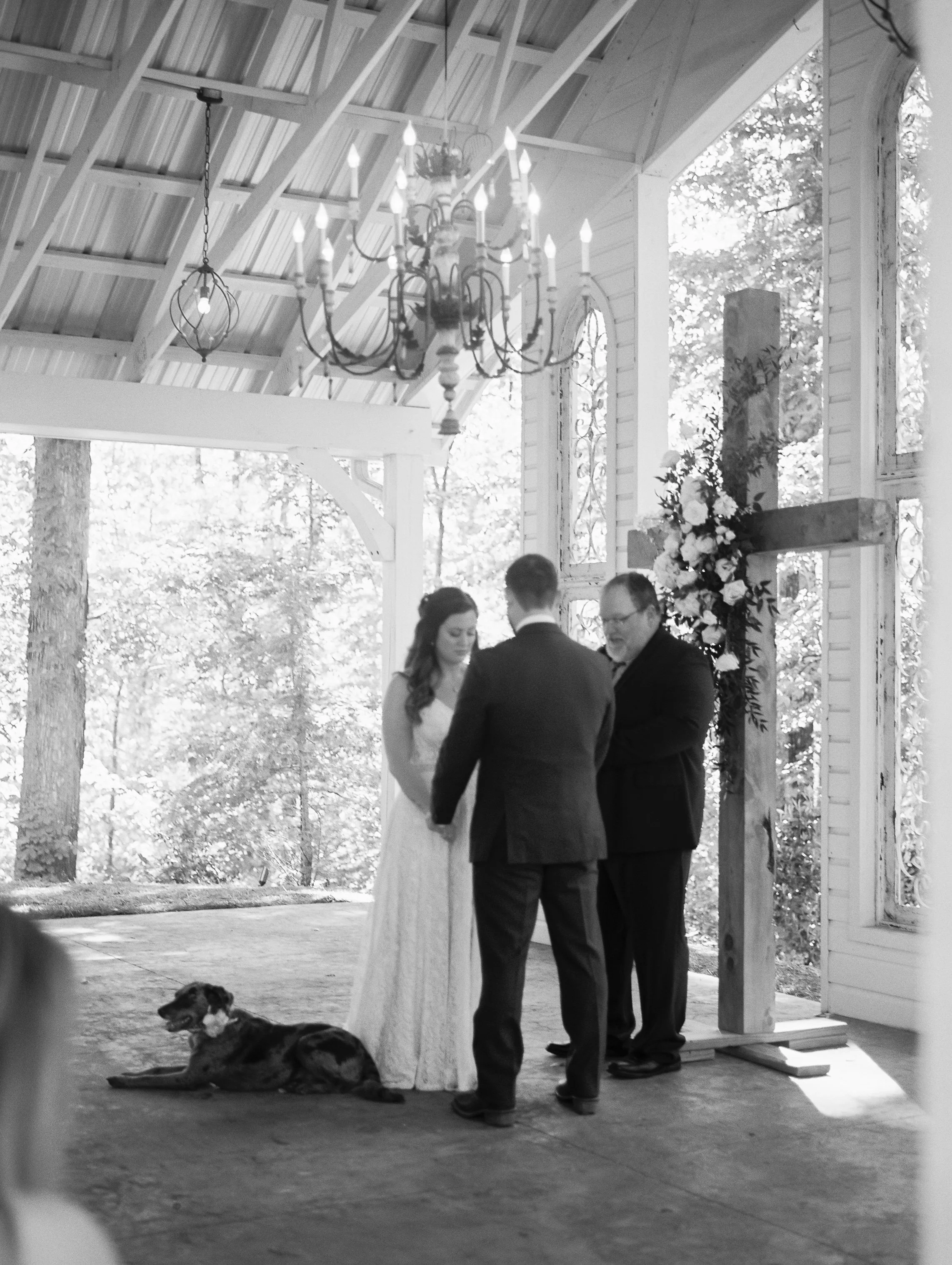 Black and white photo of the bride and groom holding hands at the altar with the officiant, Dakota resting nearby, under a chandelier