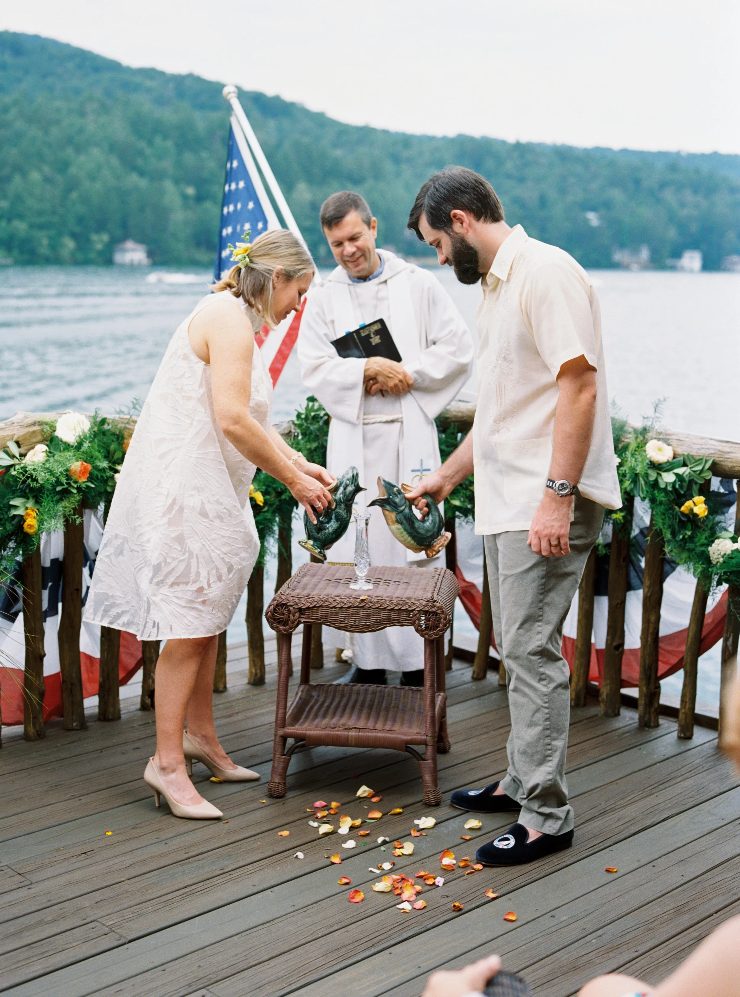 Officiant reading from a book as the bride and groom stand together during their ceremony, flower petals scattered on the deck