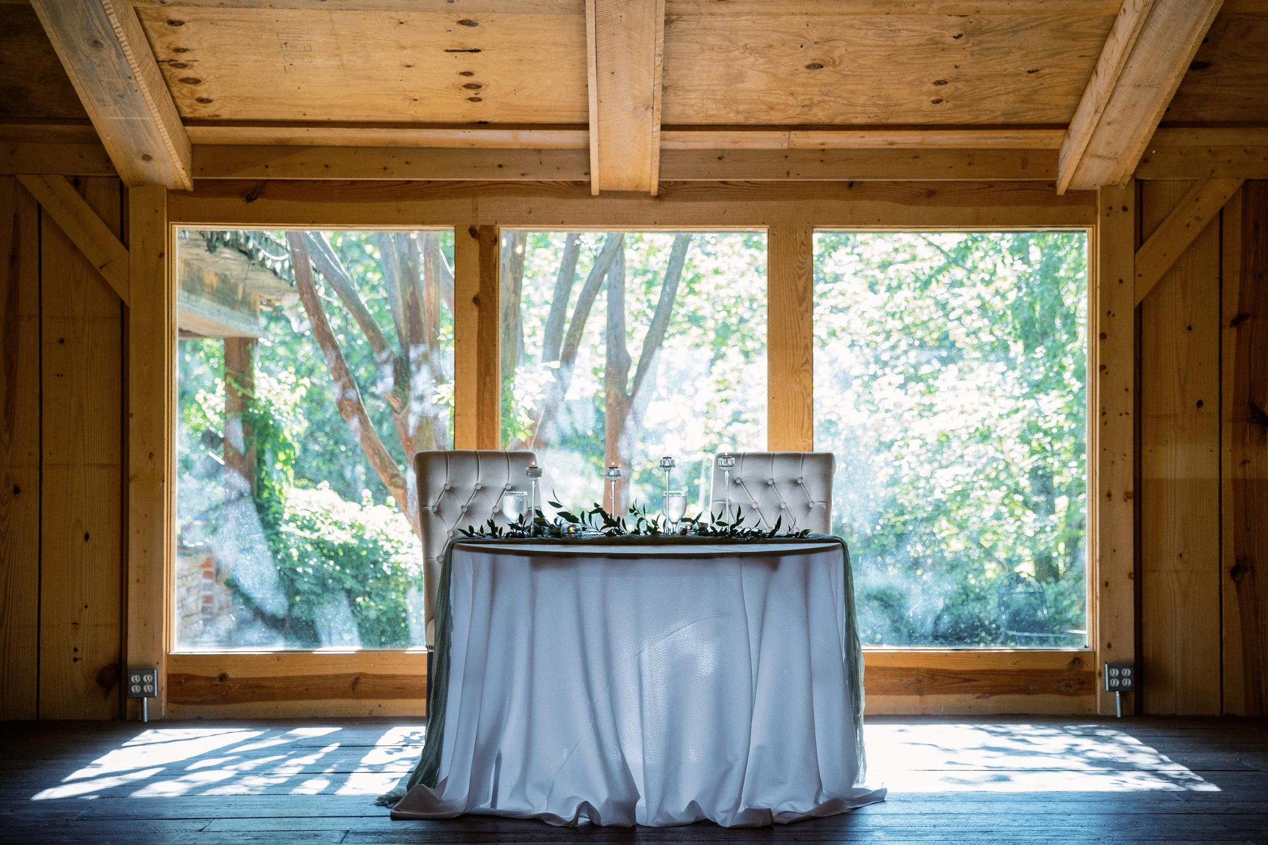 Empty sweetheart table set beneath a wood-beamed pavilion with sunlight streaming in