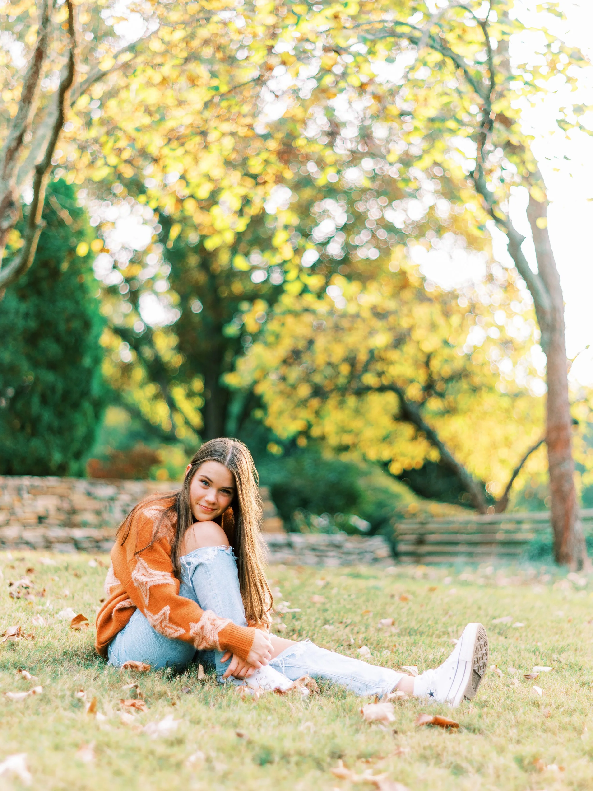 Senior portrait of Hannah sitting on the grass near a fence line, wearing an orange cardigan over a denim off-shoulder top at Kennesaw Mountain Battlefield Park in Kennesaw, Georgia