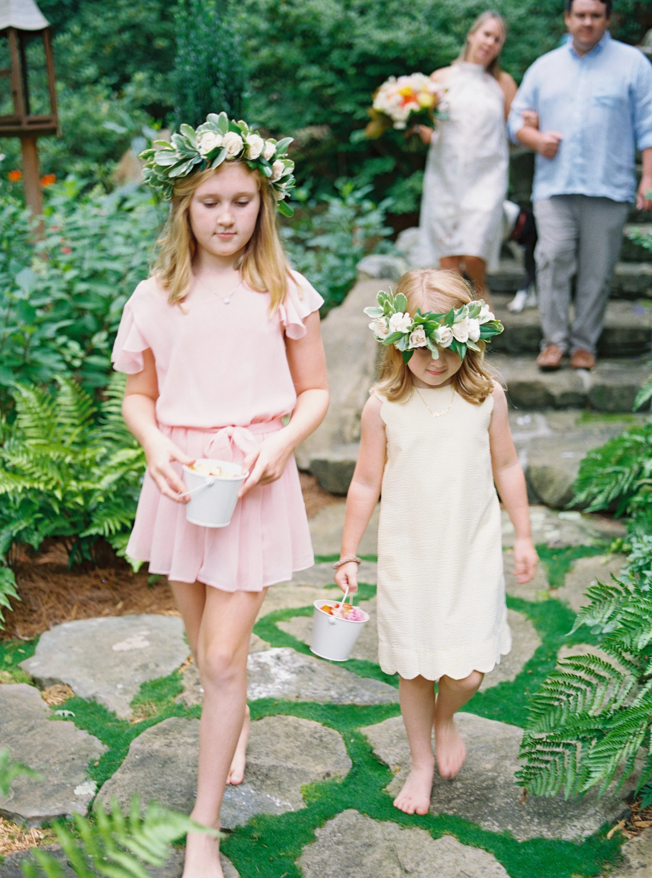 Two flower girls in pink dresses and floral crowns walking along a garden path during the wedding